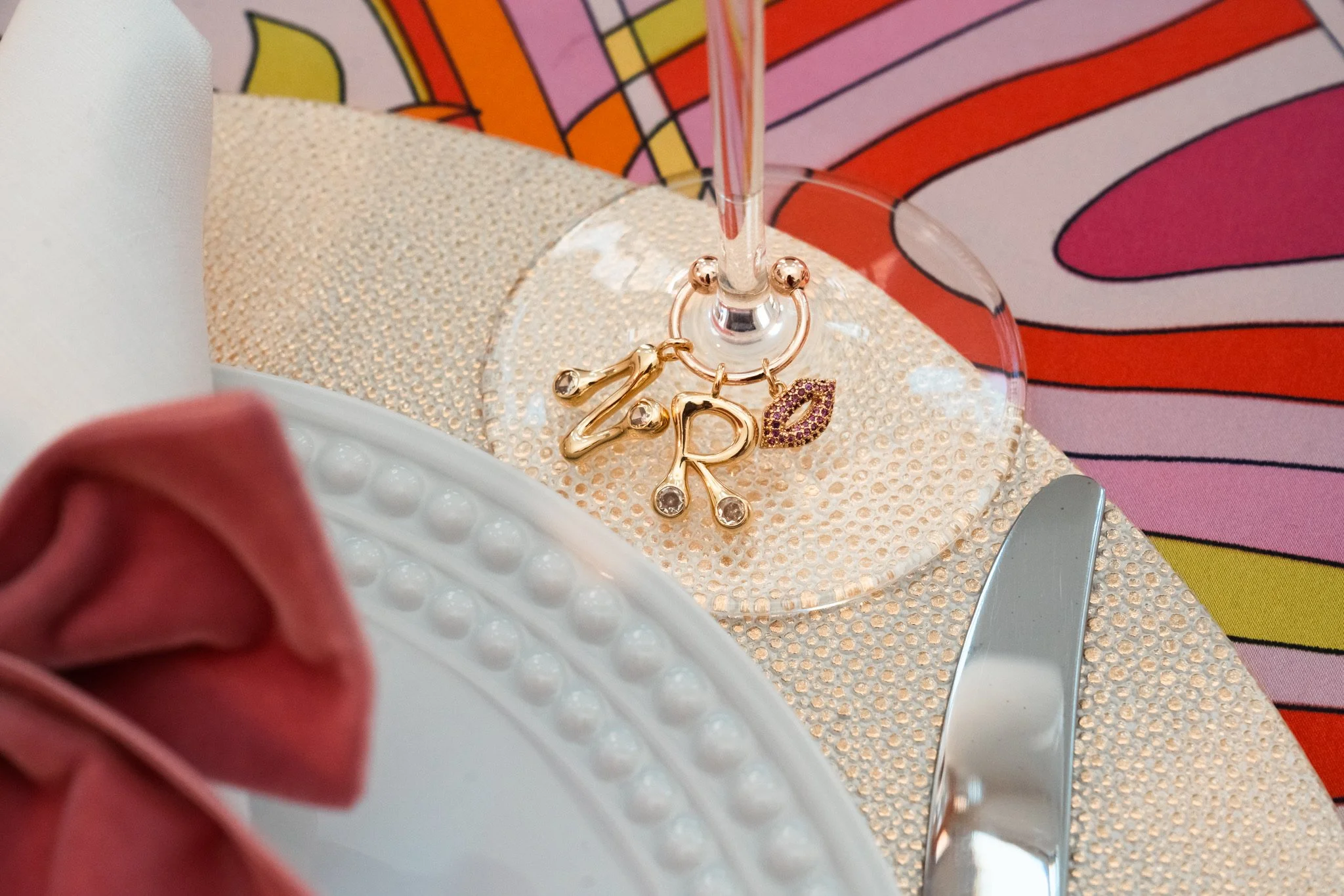 Decorative table setting with gold jewelry charms spelling 'Z R' and a pink lipstick kiss charm on a glass stem on a golden textured placemat, with a colorful background and a pink fabric napkin.