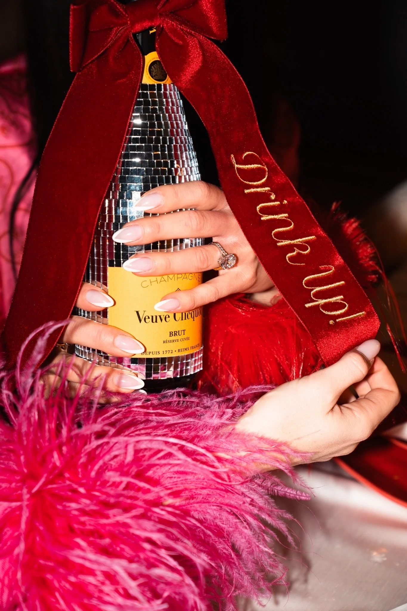 A person holding a Veuve Clicquot champagne bottle wrapped in a mosaic-style cover, with a red velvet ribbon that says 'Drink Up!' around their neck, and wearing a pink feathered garment.