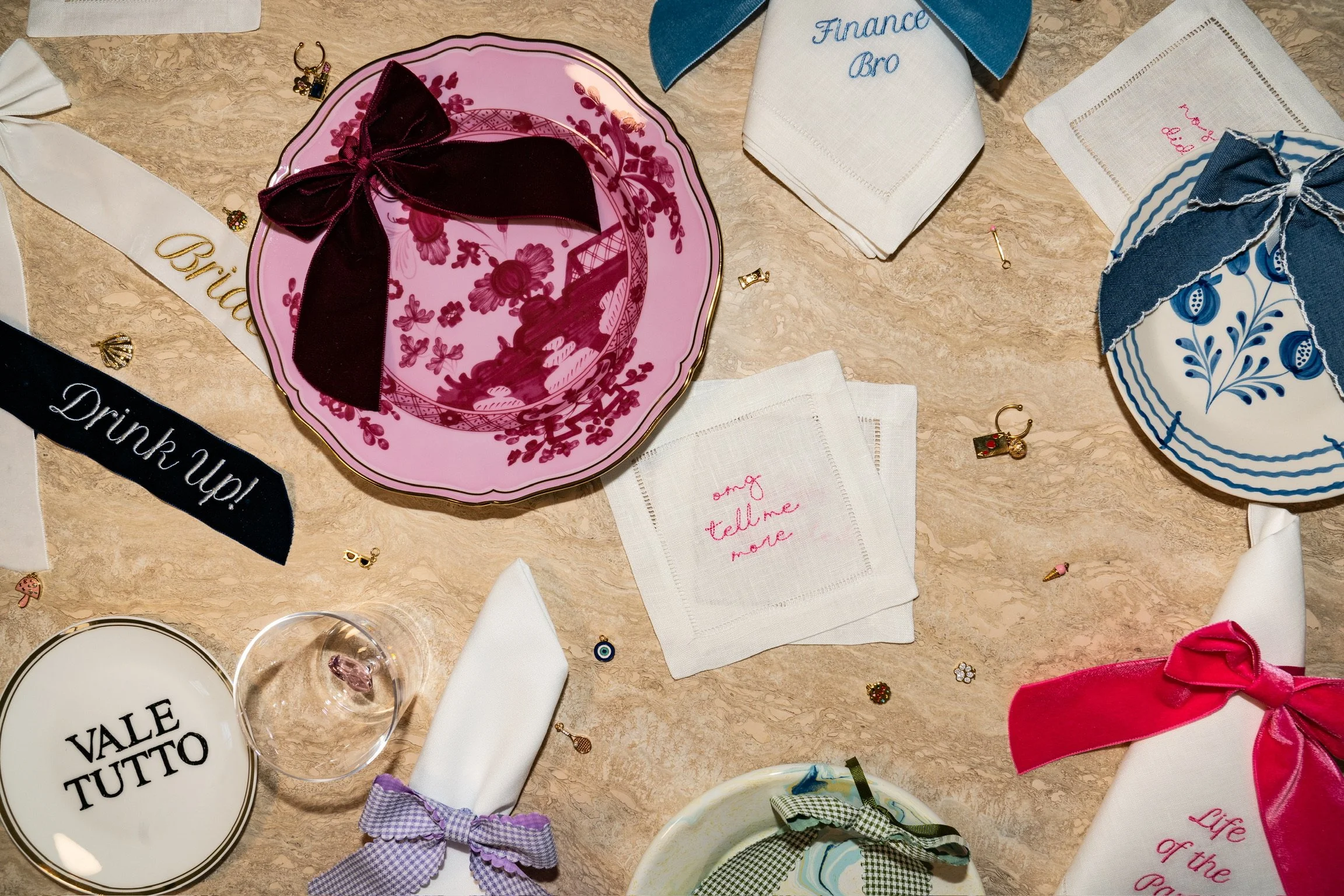 Decorative table setting with embroidered napkins, colorful plates, ribbons, and small jewelry charms for a celebratory event.