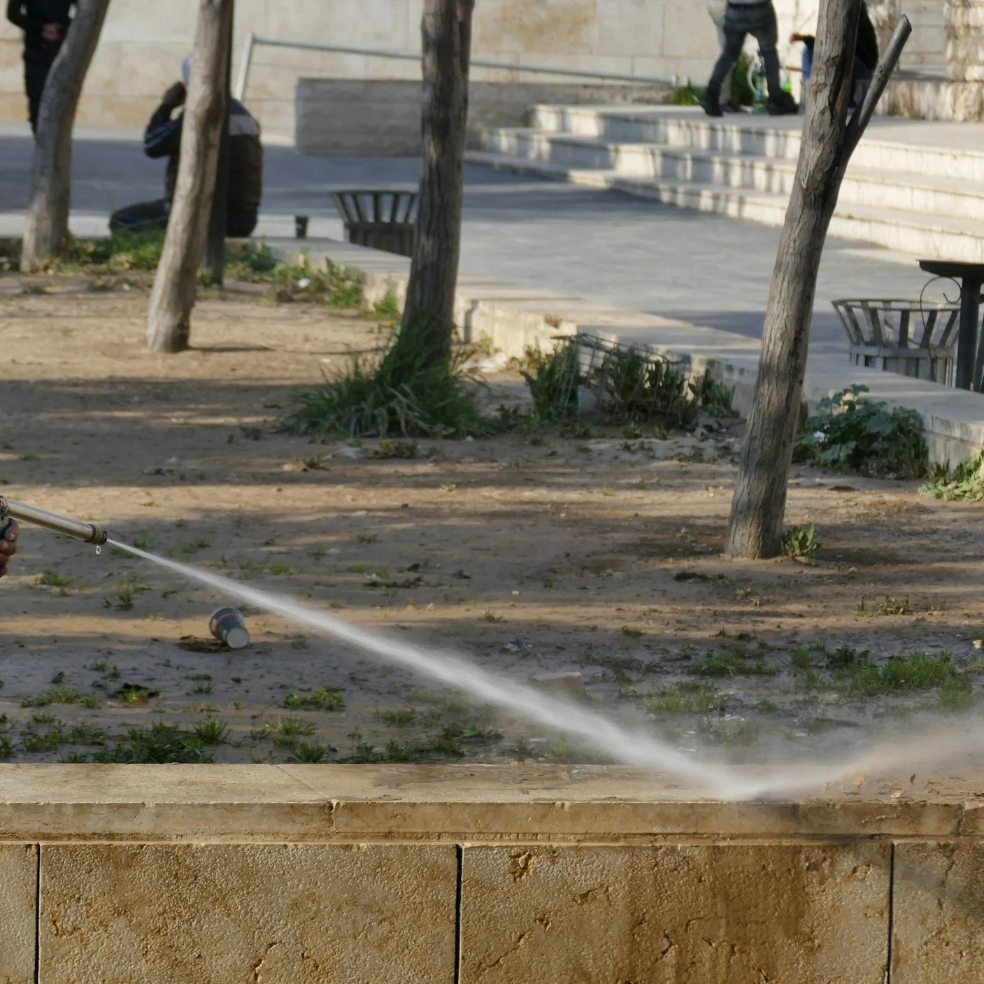 A person watering plants with a garden hose in an outdoor park or courtyard area with trees, benches, a trash can, and a set of stairs in the background.