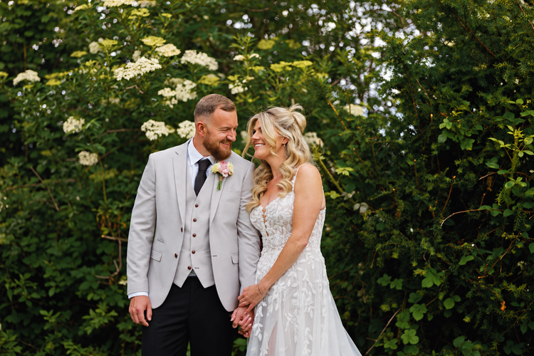 Bride and groom smiling beside a flowering hedge