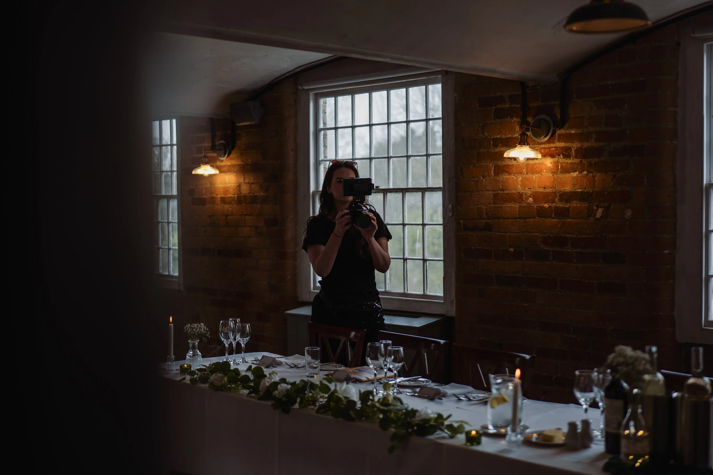 A woman taking a photo with a camera in a dimly lit rustic dining room with a brick wall and large windows, decorated with candles and a long dining table.