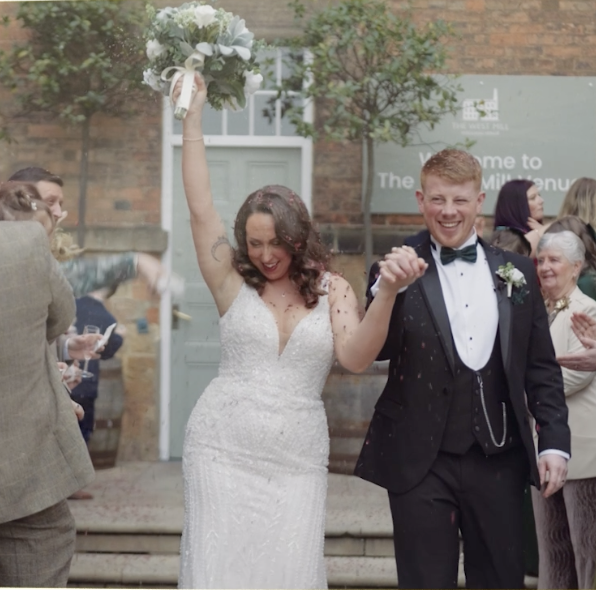 Bride and groom celebrating their wedding amidst guests, with the bride holding a bouquet of white flowers up in the air, smiling.