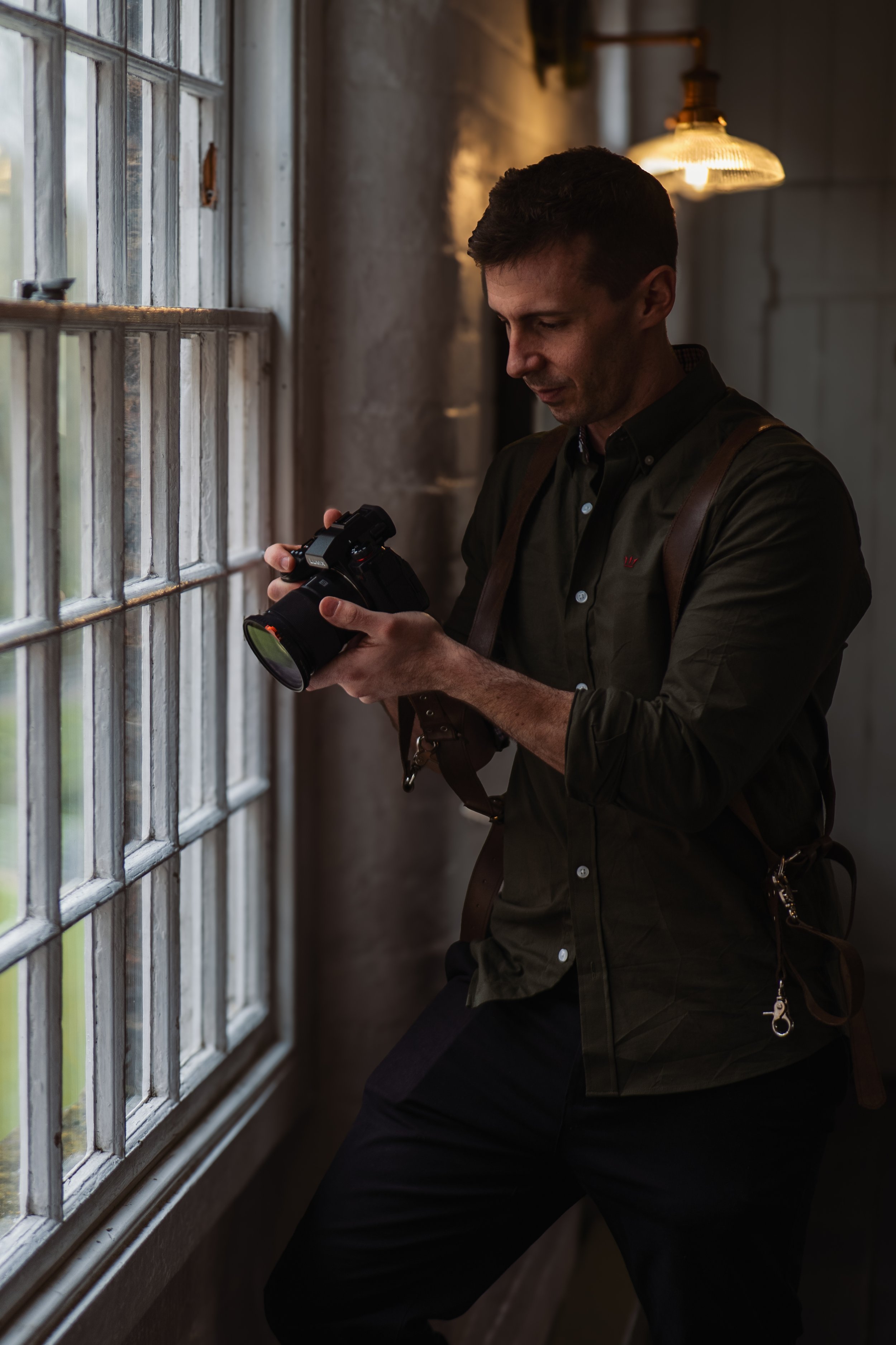 A man wearing a dark green button-up shirt and black pants is standing by a window, holding a camera and looking at it. The setting appears to be indoors with a warm light above.