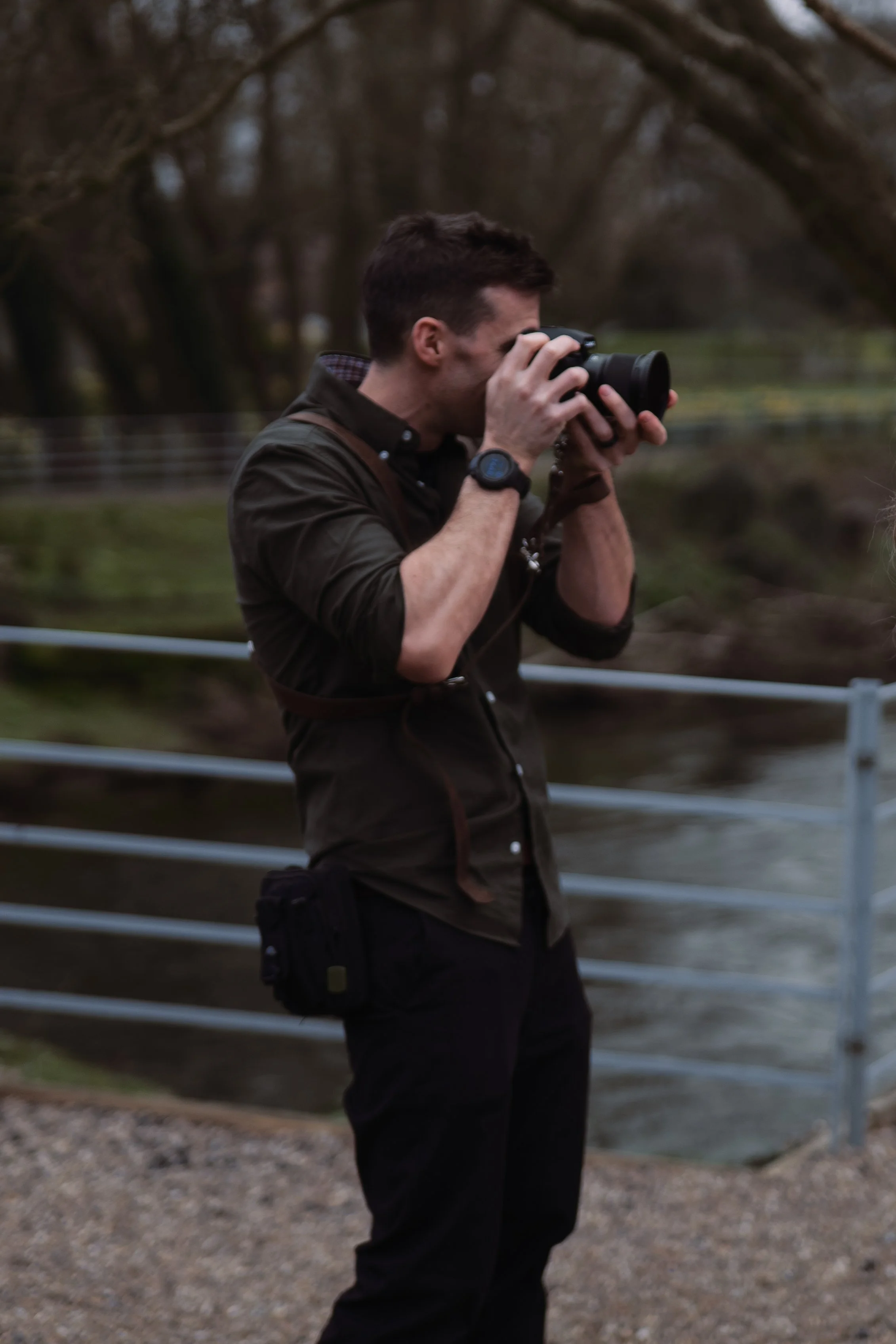 A man taking a photograph with a camera outdoors near a body of water and trees.