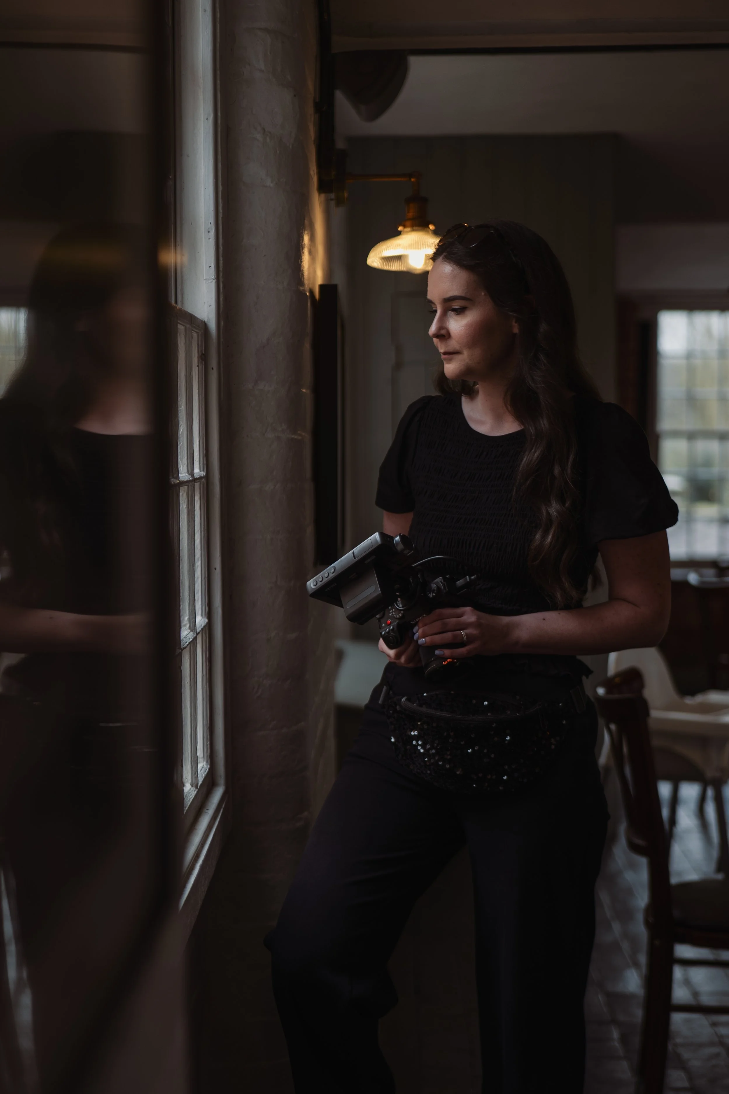 A woman with long dark hair, wearing a black top and pants, holding a video camera, standing indoors near a window.