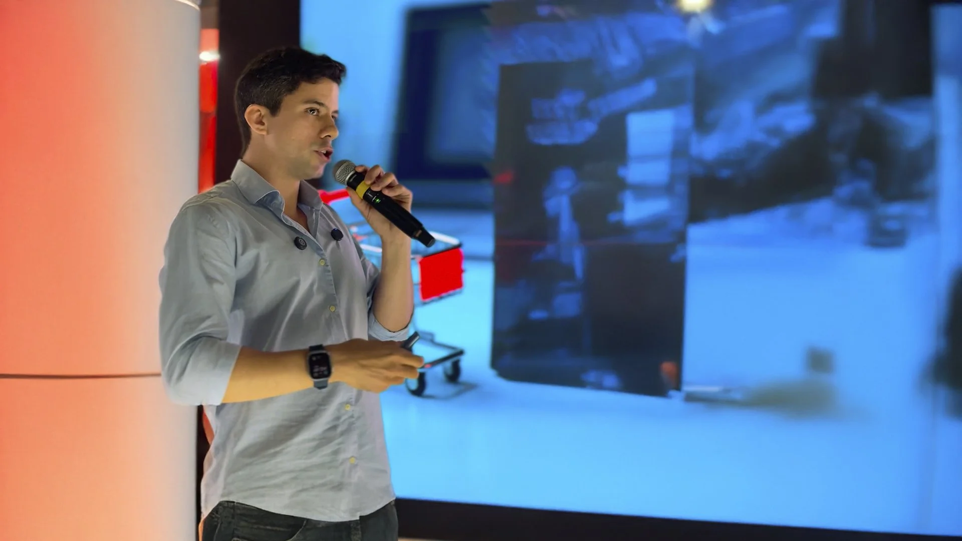 Homem apresentando com microfone em frente a uma tela grande com imagens desfocadas, em uma sala de apresentação.