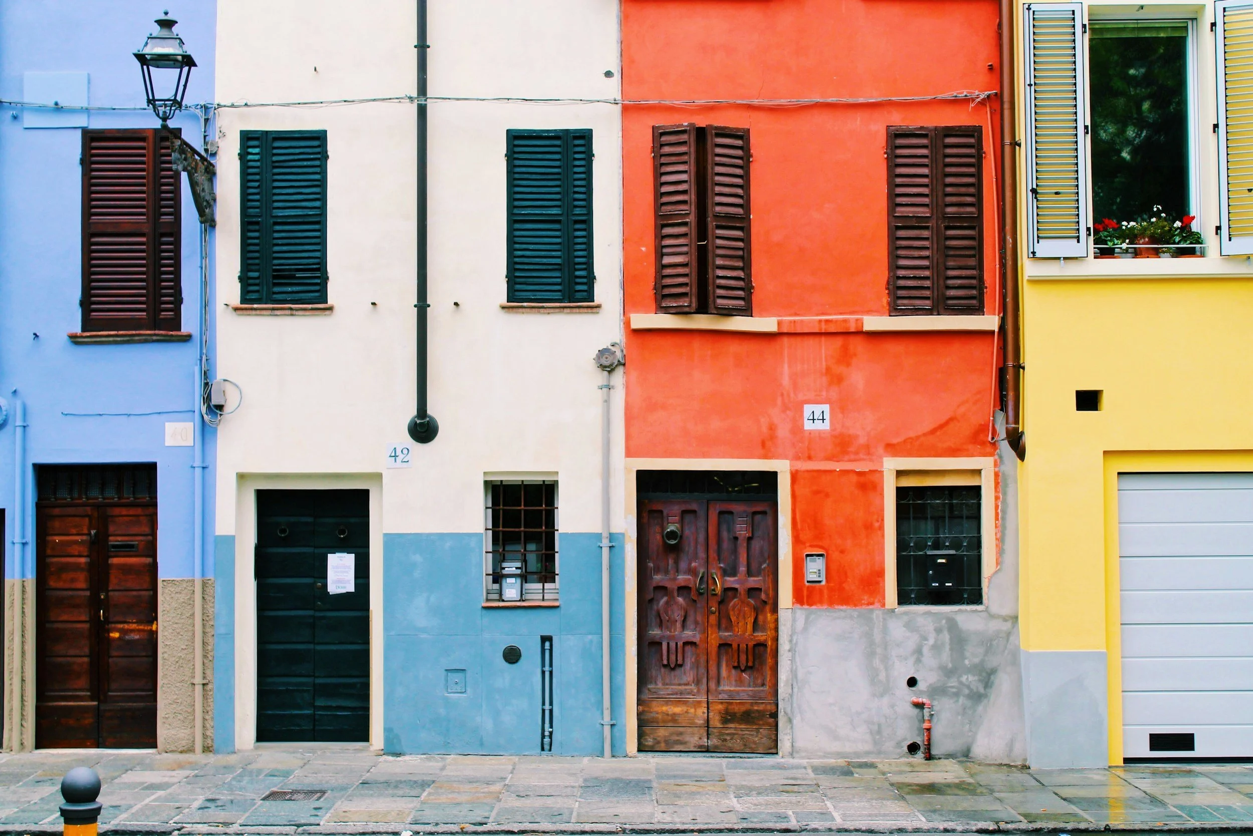 Colorful row of houses with closed shutters, doors, and small balconies on a cobblestone street, featuring blue, white, orange, and yellow facades.