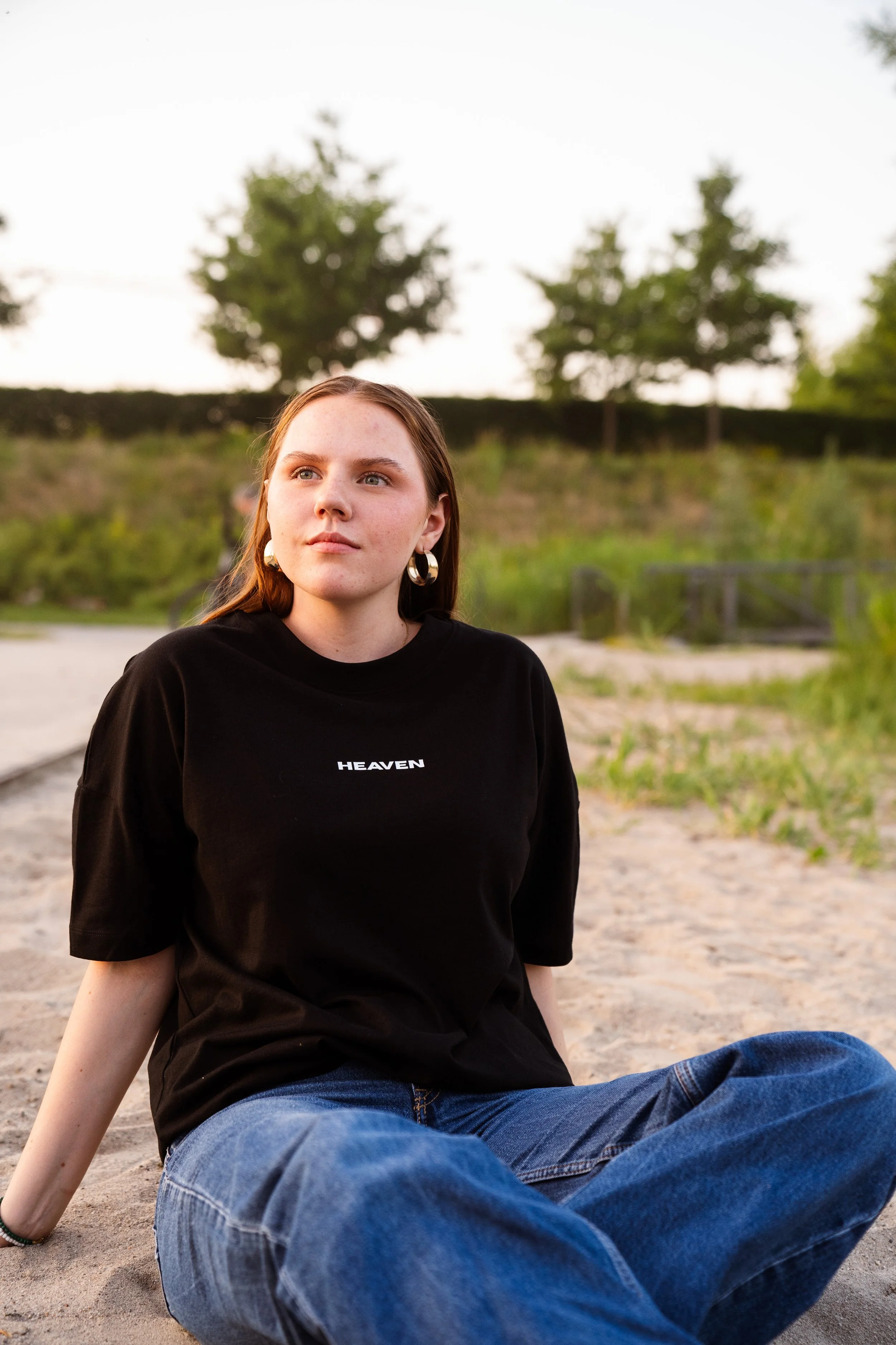 Junge Frau sitzt am Sandstrand, trägt ein schwarzes T-Shirt mit 'HEAVEN' geschrieben, trägt große Ohrringe, hat lange braune Haare, sieht nachdenklich in die Ferne, im Hintergrund Bäume und grüne Landschaft bei Sonnenuntergang.