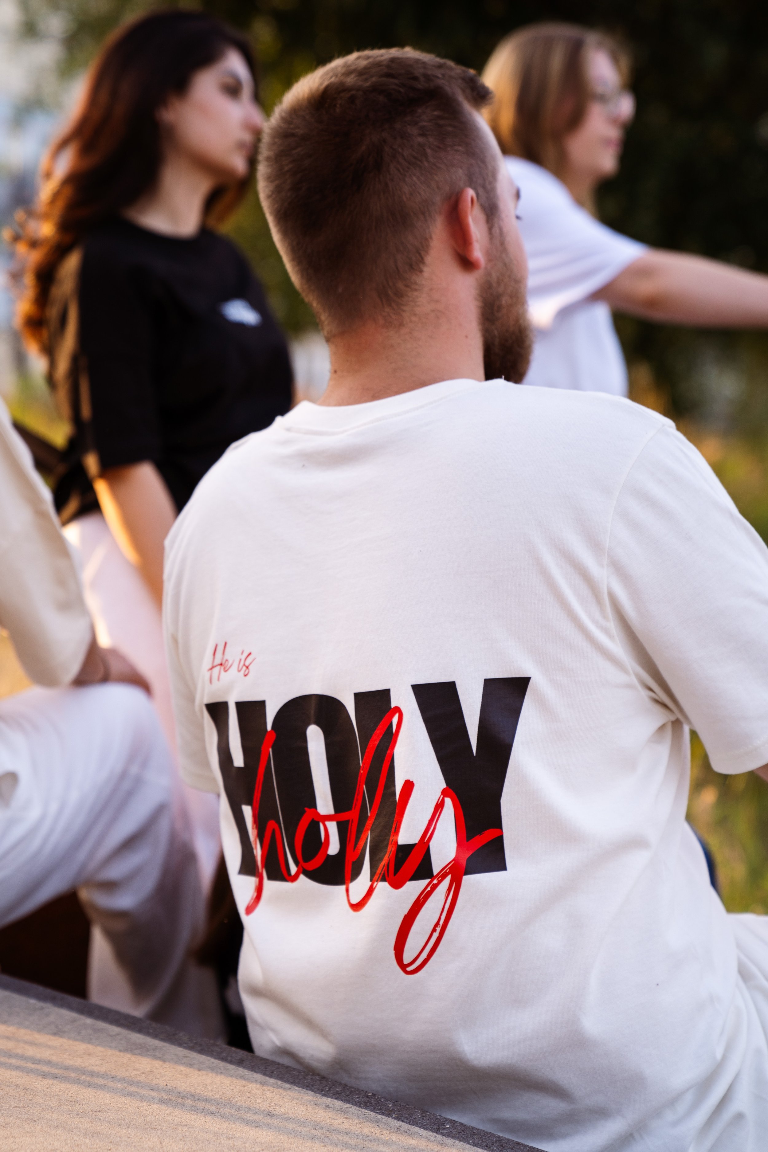 Junge Männer und Frauen sitzen im Freien, sitzen auf einer Bank. Der Fokus liegt auf einem Mann mit Natural gefärbten T-Shirt, auf dem in schwarz und rot "Holy" steht