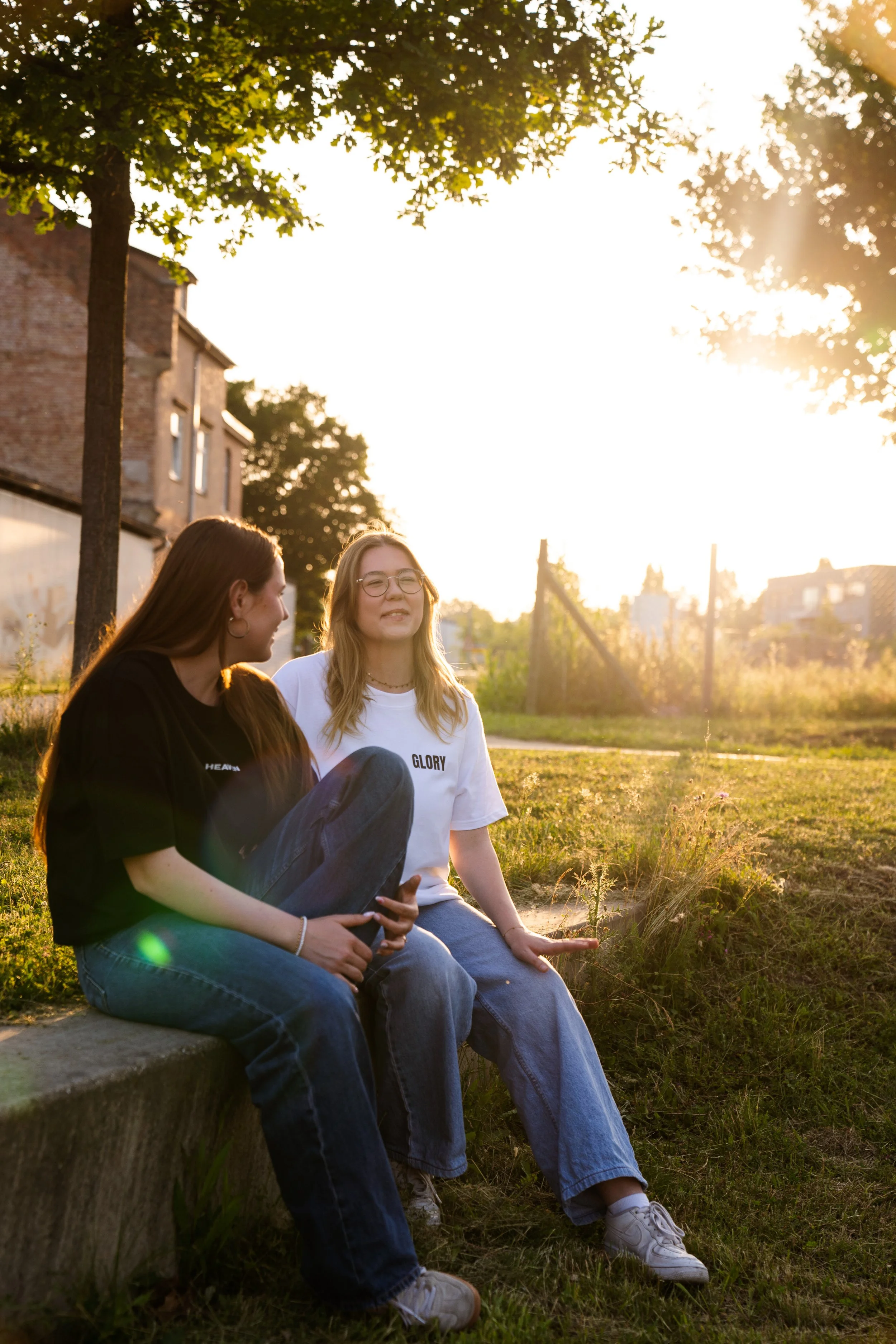 Zwei junge Frauen sitzen auf einer Mauer im Sonnenuntergang in einem Garten, lachen und unterhalten sich.
