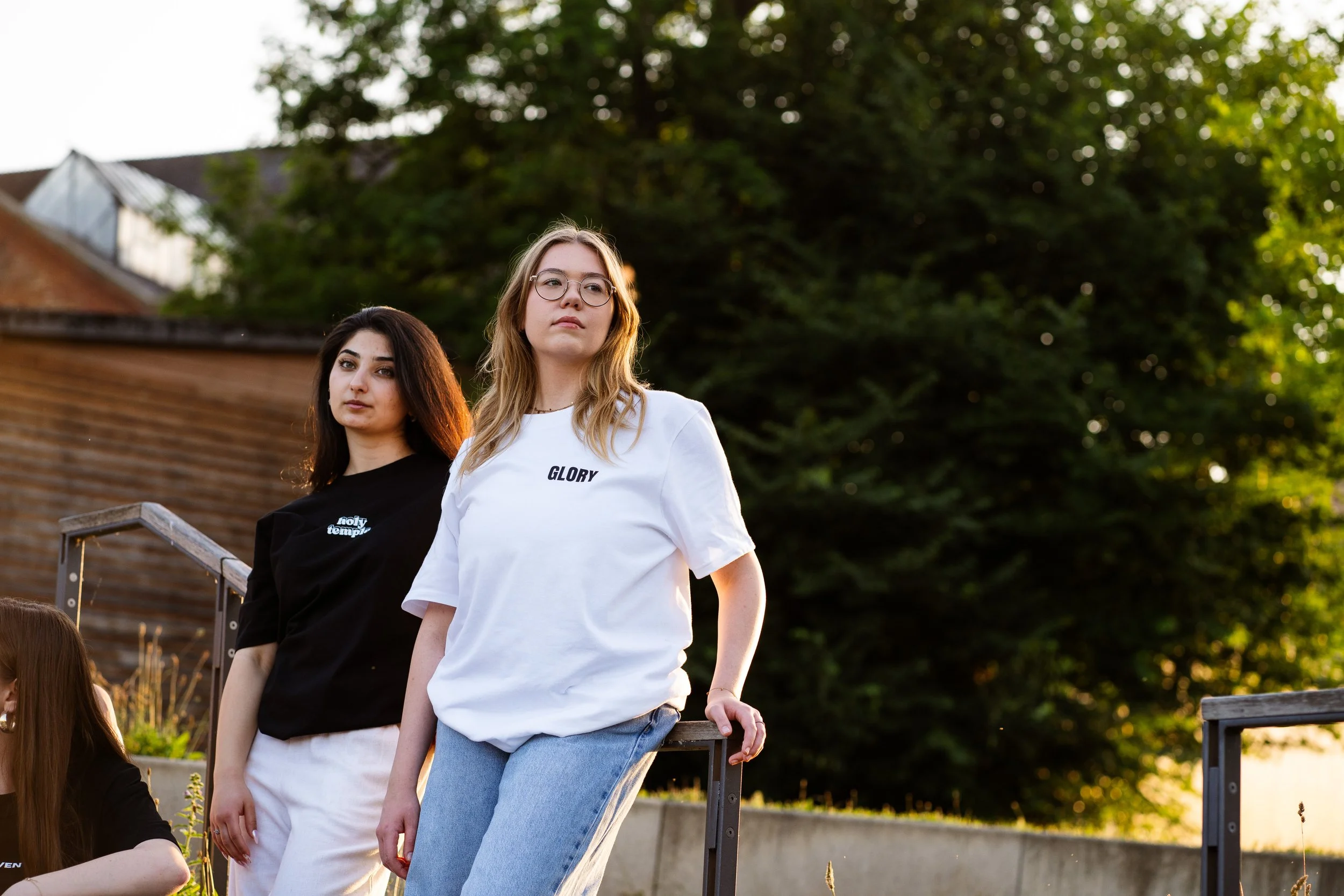 Zwei junge Frauen stehen im Freien, eine trägt ein schwarzes T-Shirt mit weißem Text, die andere ein weißes T-Shirt mit dem Wort 'GLORY'. Es ist Abend und die beiden schauen nach vorne, während im Hintergrund Bäume und ein Gebäude zu sehen sind.