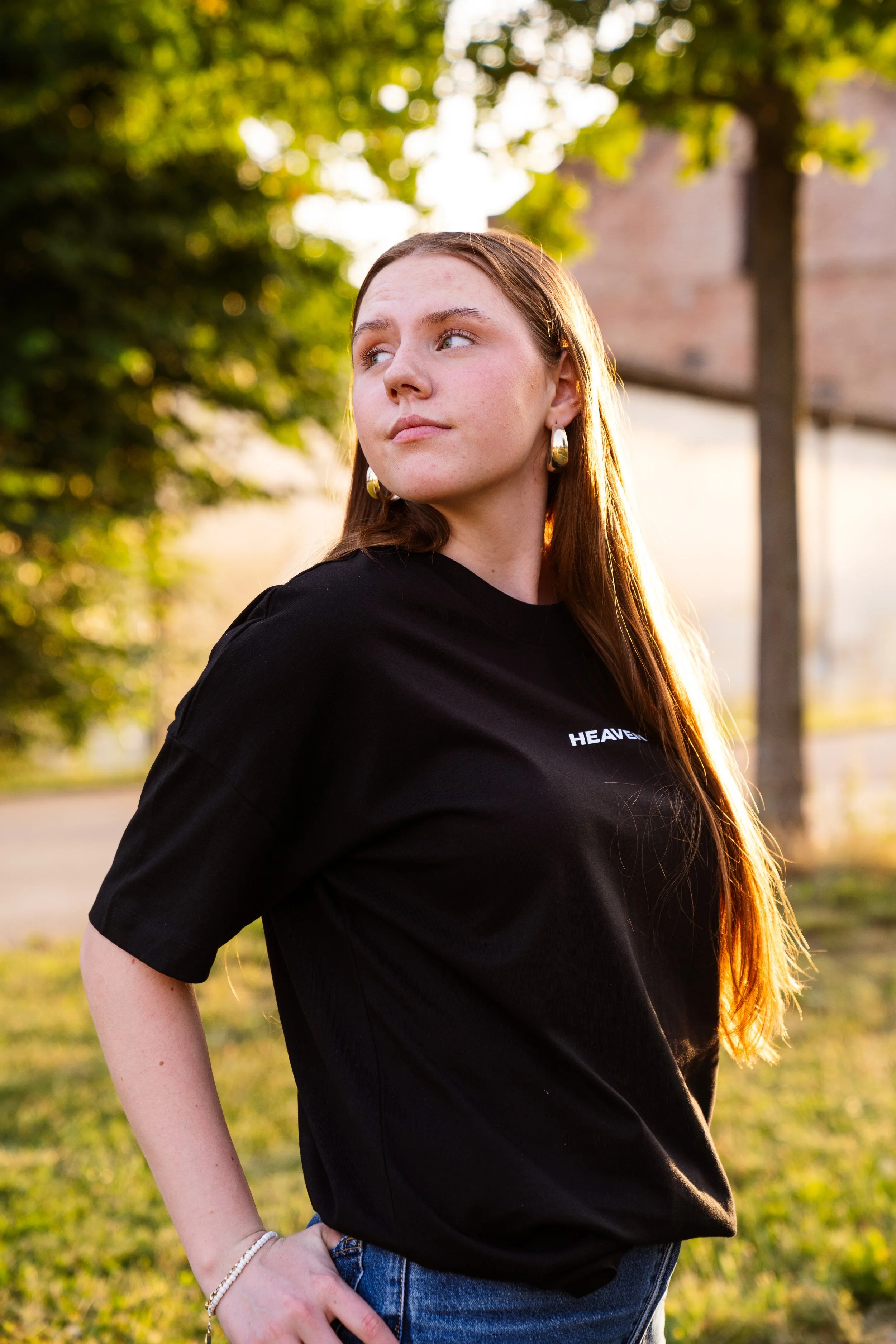 Junge Frau mit langen roten Haaren, schwarzen T-Shirt mit Aufschrift "Heaven" steht draußen im Sonnenlicht, neben einem Baum, mit Blick nach rechts, im Hintergrund ein Gebäude.