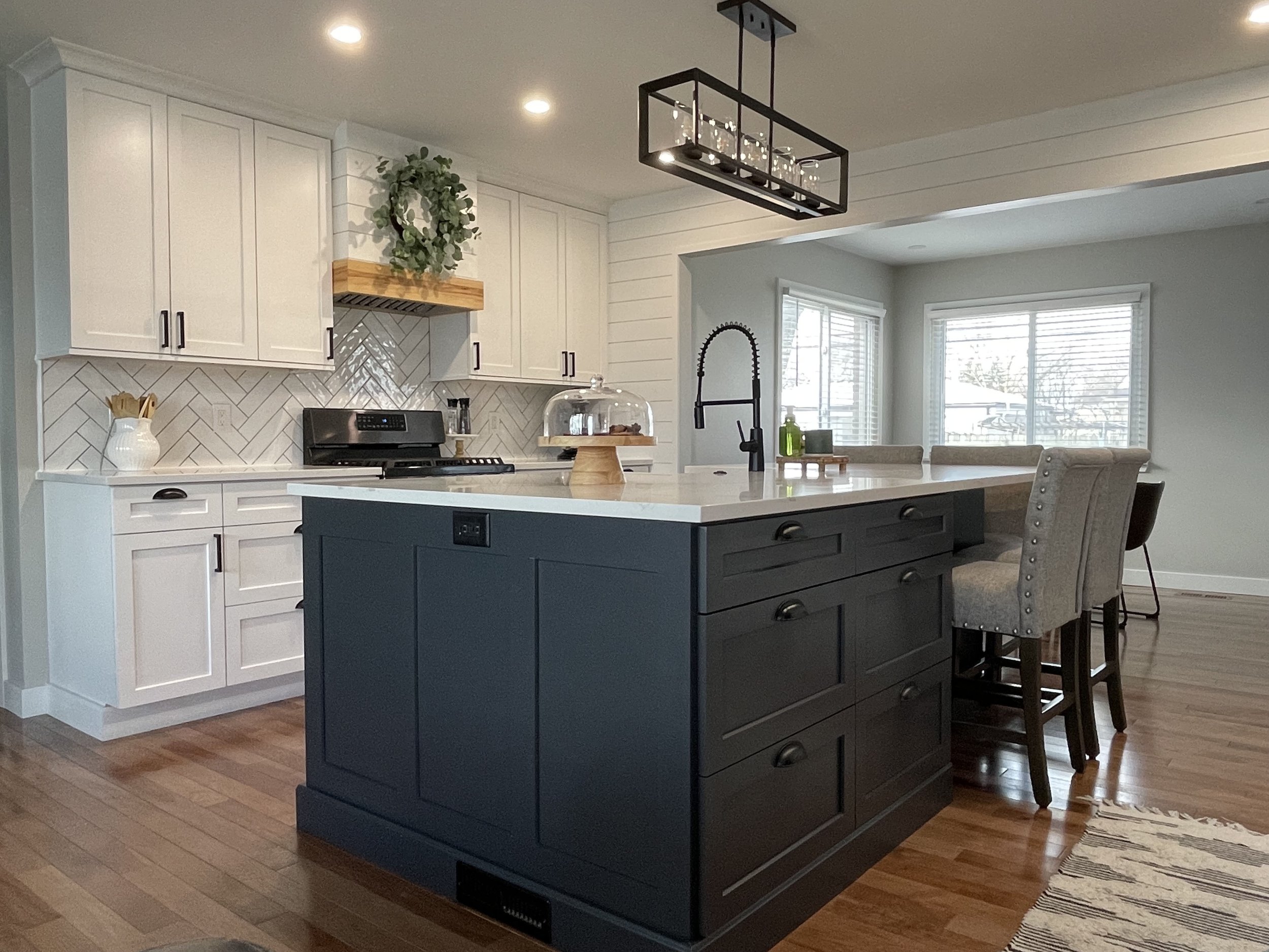 Modern kitchen with white upper cabinets, a black stove, a blue island with drawers, beige upholstered chairs, and large windows with white blinds.