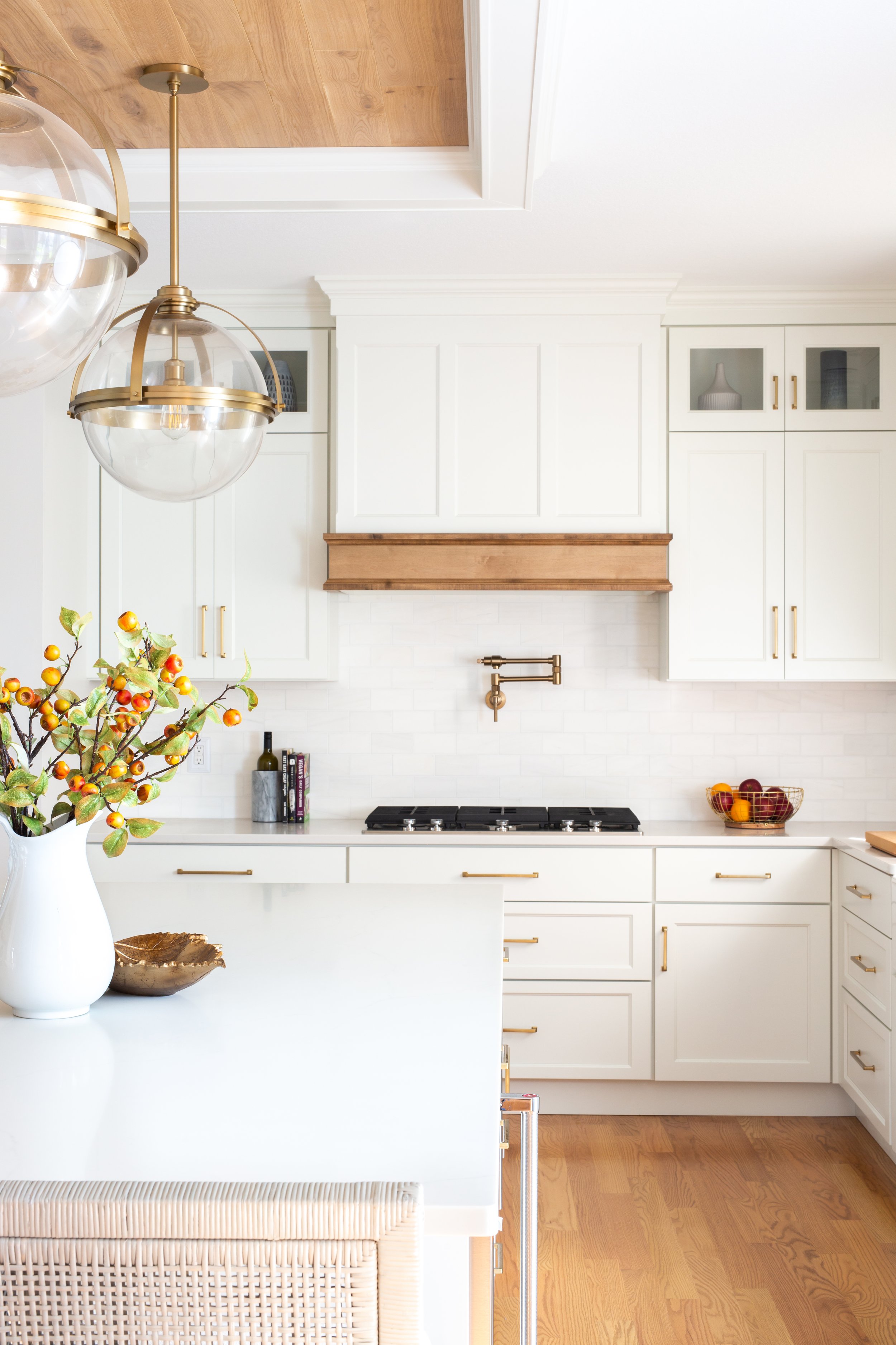 A bright white kitchen with modern cabinetry, a gas stove, and a wooden range hood. There are decorative items including a bowl of fruit, a book, and a vase with branches on the countertop, with warm wood flooring and two hanging glass globe light fi