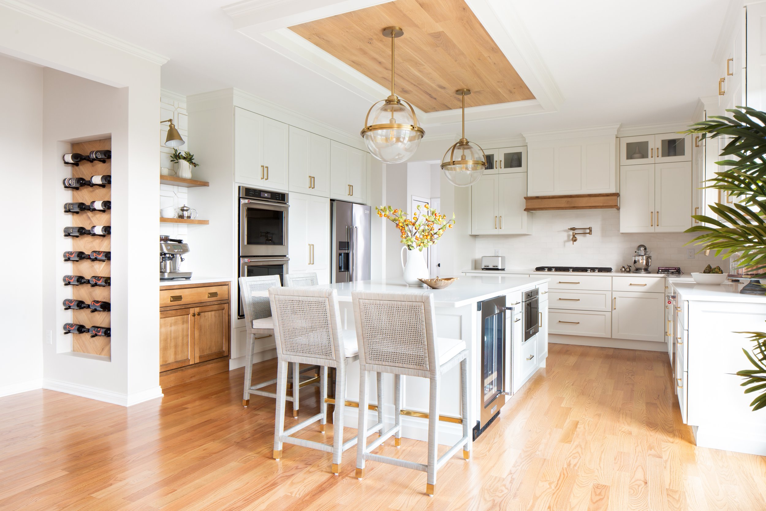 Bright, modern kitchen with white cabinets, wooden accents, a central island with a white countertop, bar stools, and built-in appliances, including a refrigerator, oven, and microwave. Decor includes a large floral vase, a bowl, a coffee machine, an