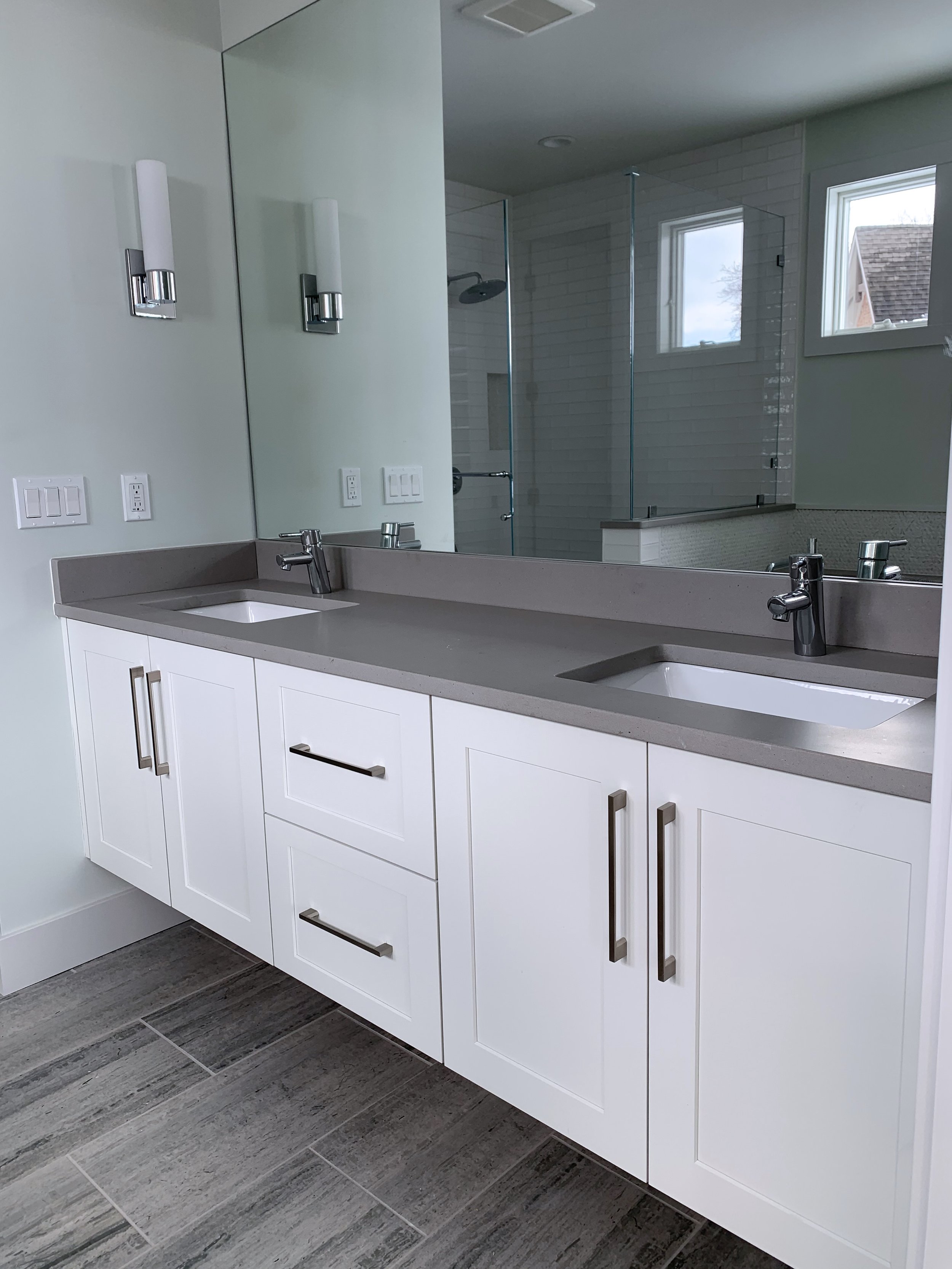 Modern bathroom vanity with two sinks and white cabinets, large mirror, and gray countertop, with a shower and windows reflected in the mirror.