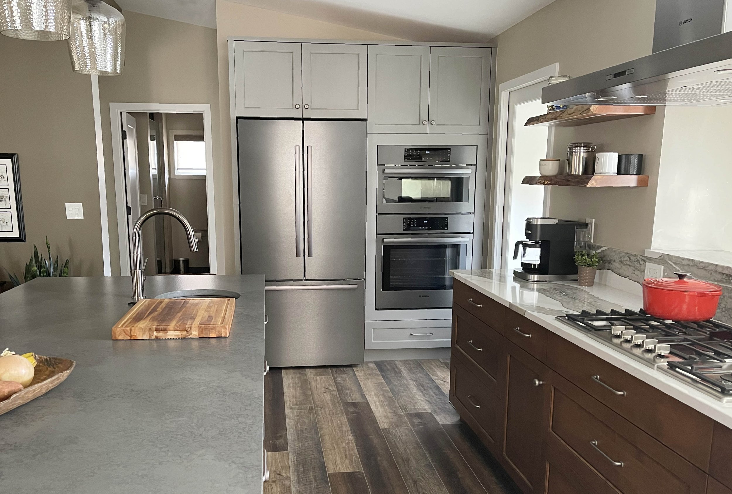 Modern kitchen with stainless steel refrigerator, built-in oven, microwave, and gas stove. Brown wooden cabinets, grey island counter with a cutting board, and open floating shelves with kitchenware. Wooden flooring and a small window.
