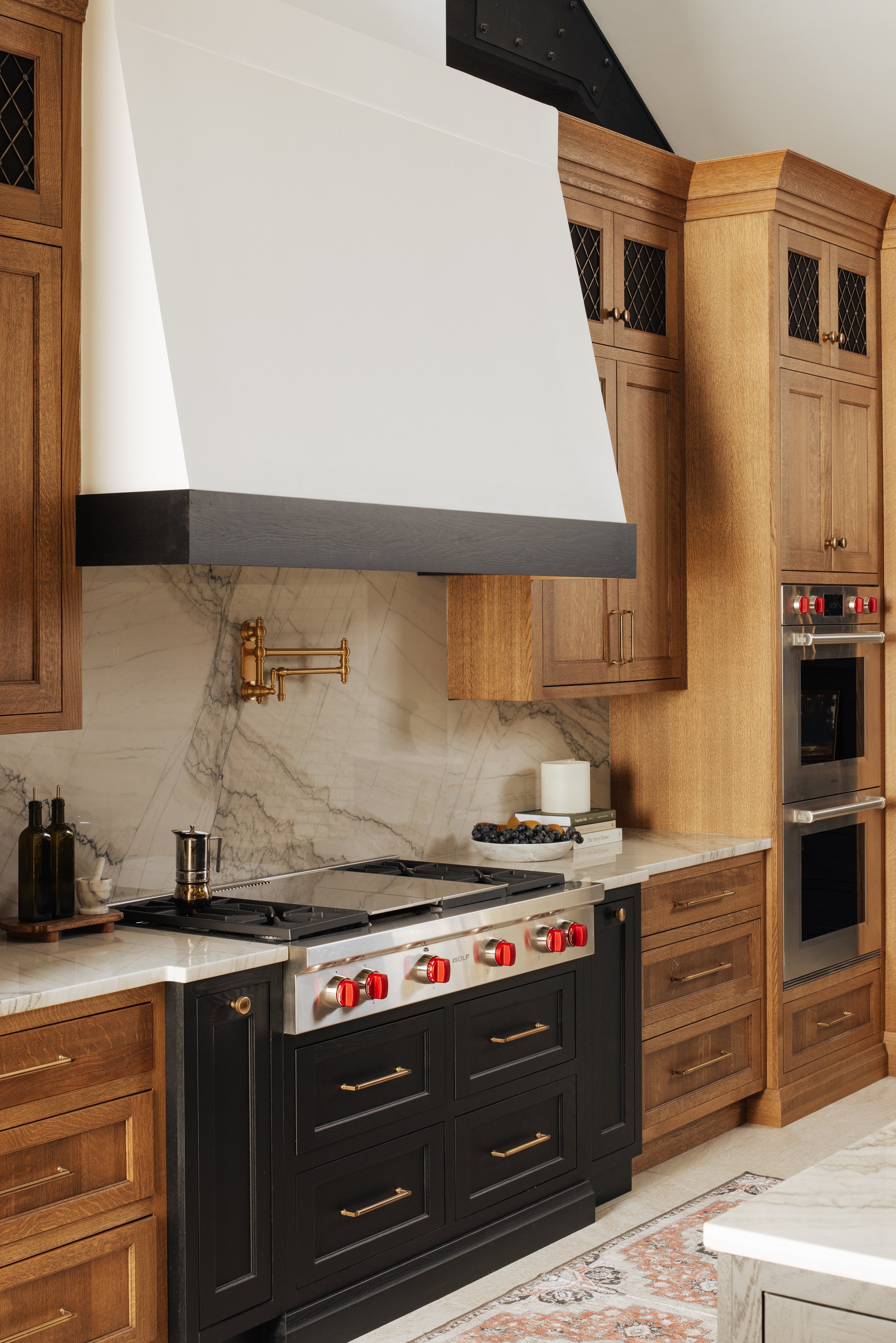 Kitchen with wooden cabinets, marble backsplash, black stove with red knobs, white range hood, and a built-in oven.