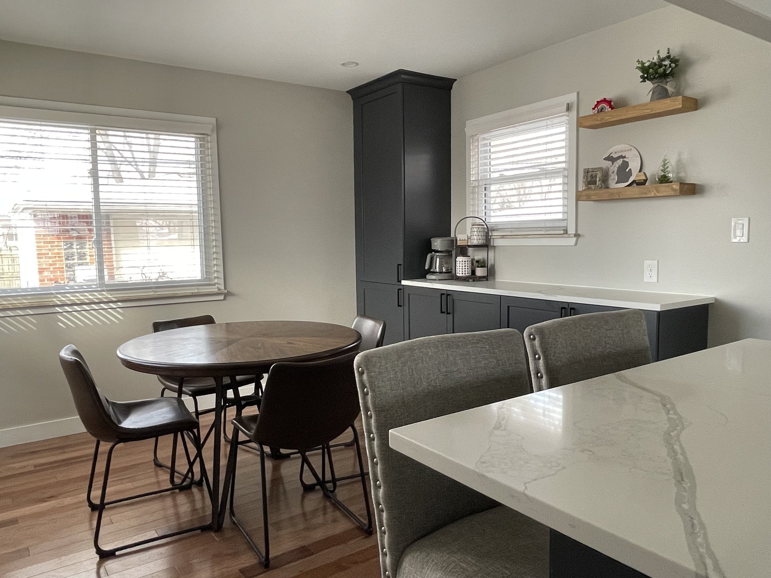 Dining area with a round wooden table and four black chairs, gray cabinetry with a coffee maker, and decorative shelves on a white wall, with windows letting in natural light and hardwood floors.