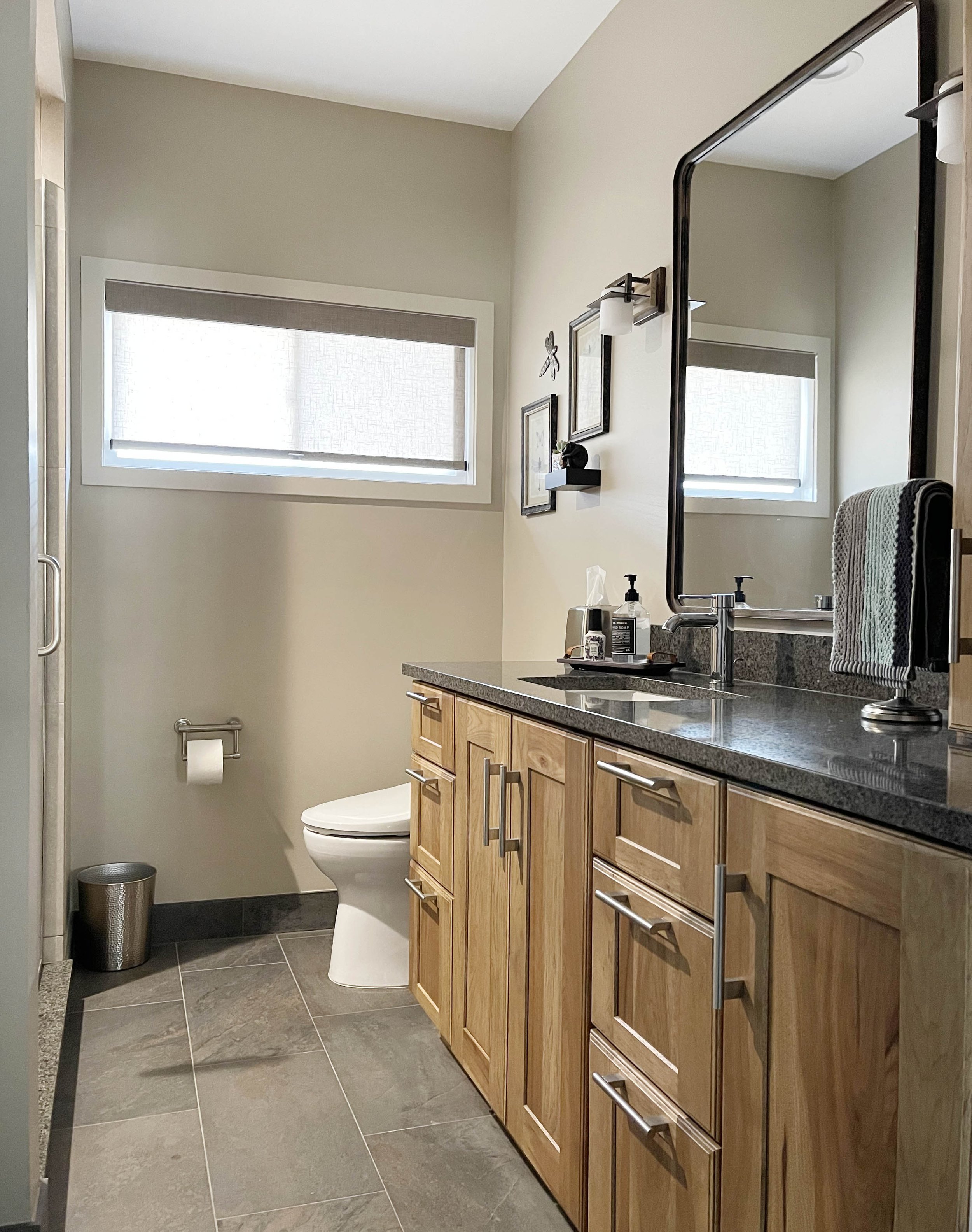 A modern bathroom with a wooden vanity, black countertop, large mirror, small window, and a toilet with a paper holder and trash can nearby.