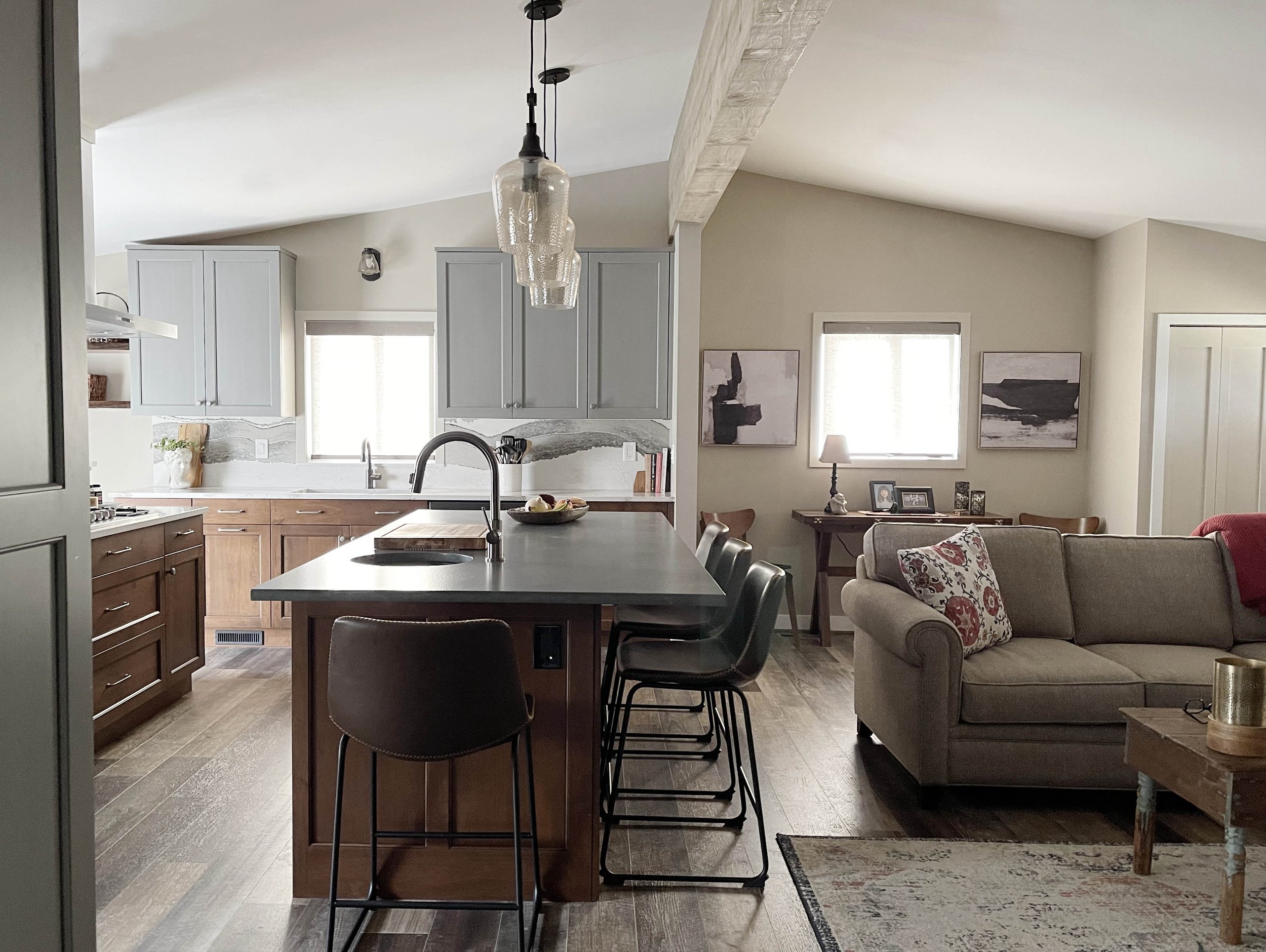 Open-concept kitchen and living room with gray upper cabinets, wood lower cabinets, a kitchen island with black barstools, beige couch with patterned pillows, and wall art.
