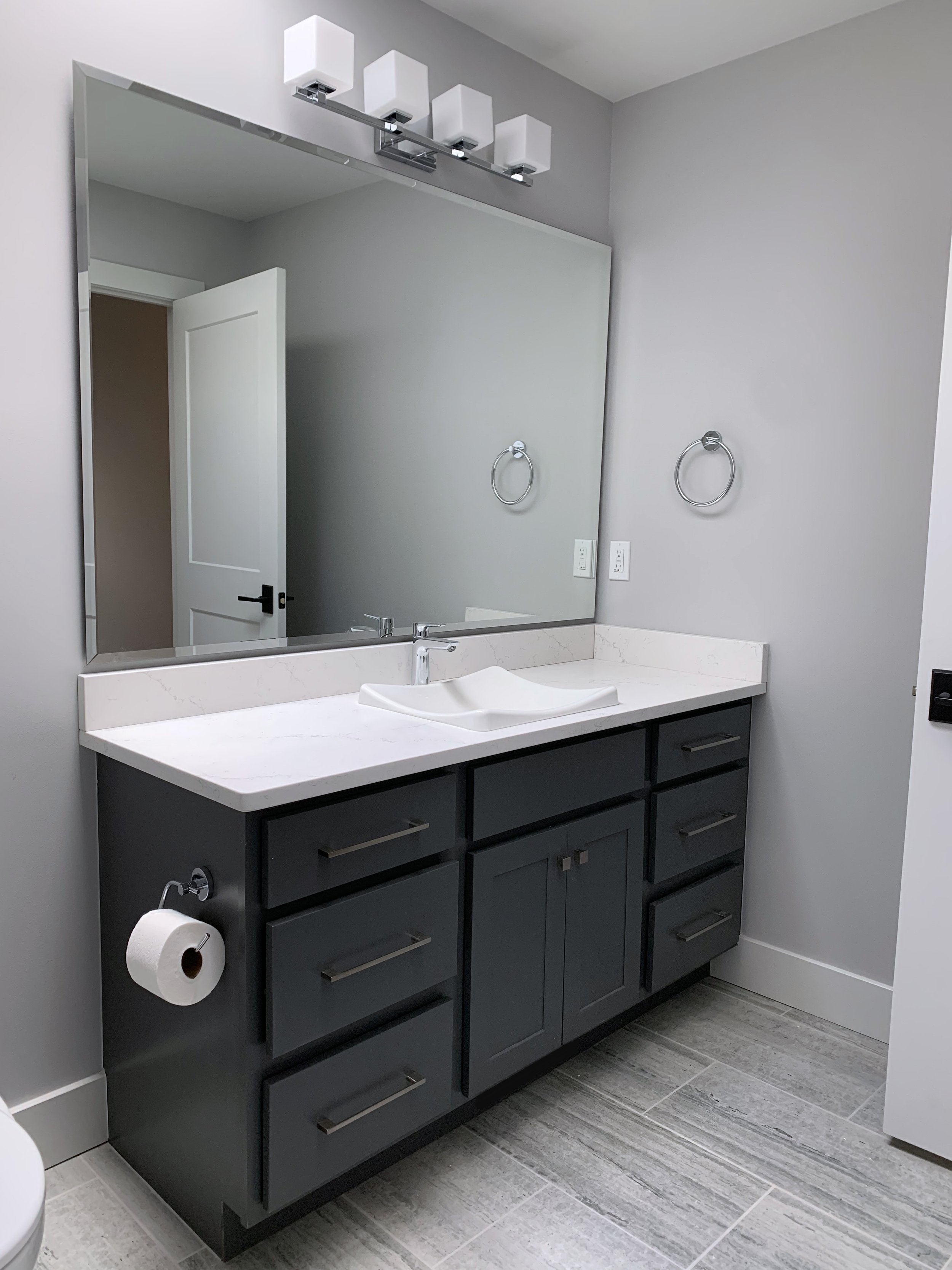 Modern bathroom vanity with a dark gray cabinet, white countertop, and white vessel sink. Large mirror above, four white light fixtures, two chrome towel rings, and a toilet paper holder on the side. Gray tiled floor and light gray walls.