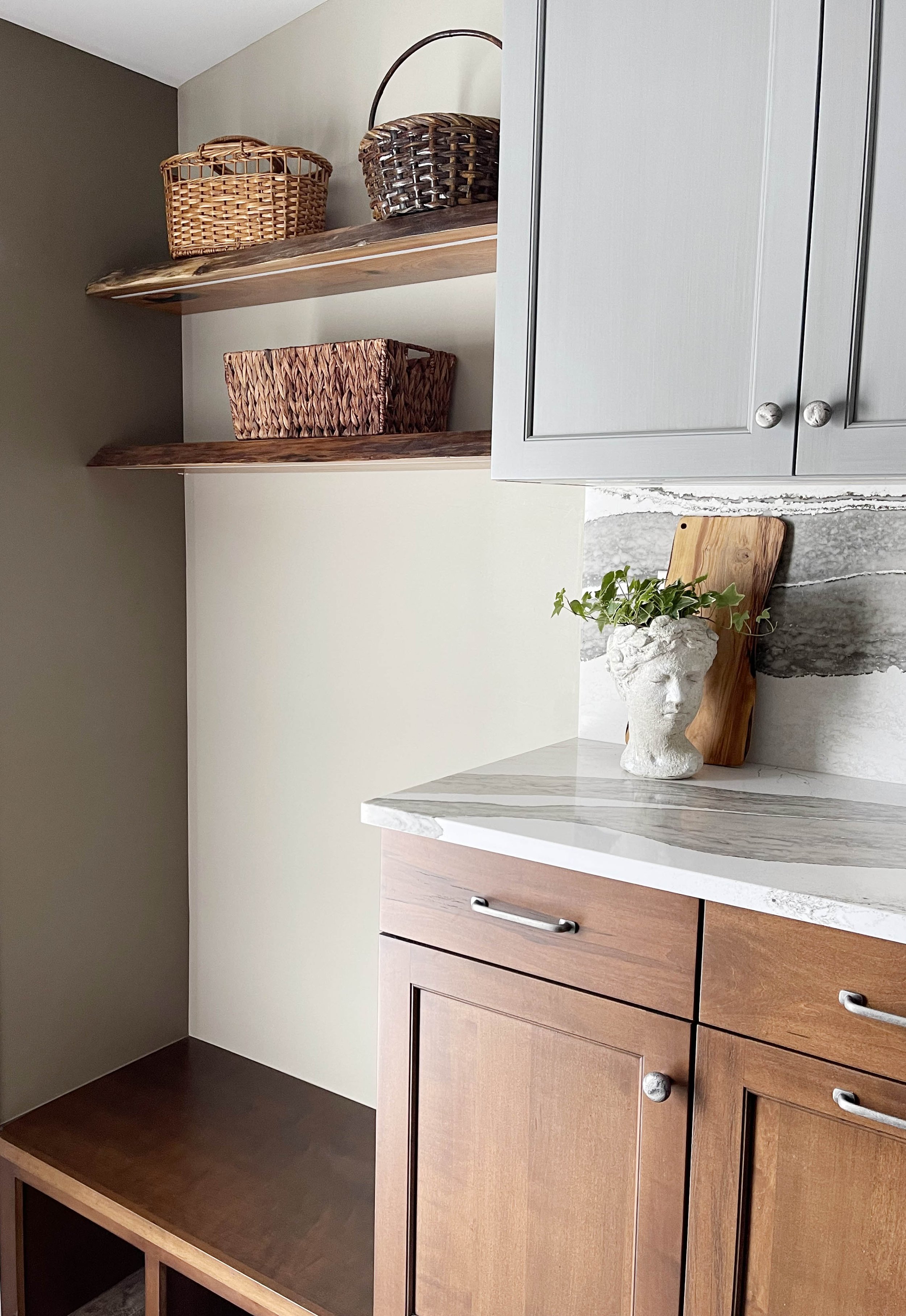 Inside a kitchen with two open wooden shelves holding baskets, a white vase with green ivy, a cutting board, gray upper cabinets, and a wooden lower cabinet.