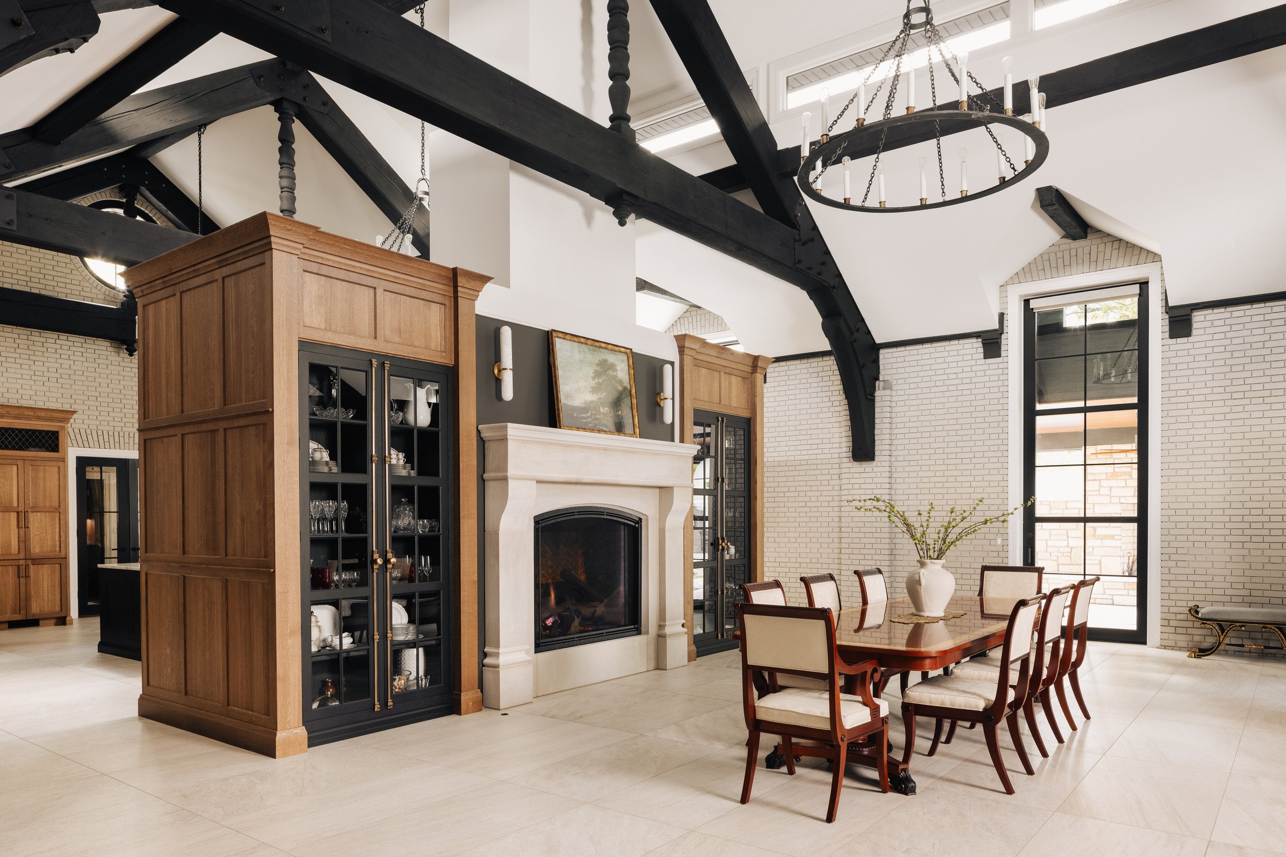 Elegant dining area with a large wooden table and eight upholstered chairs, white brick walls, a fireplace with a landscape painting above, black and wooden cabinets, large windows, and a black metal chandelier.