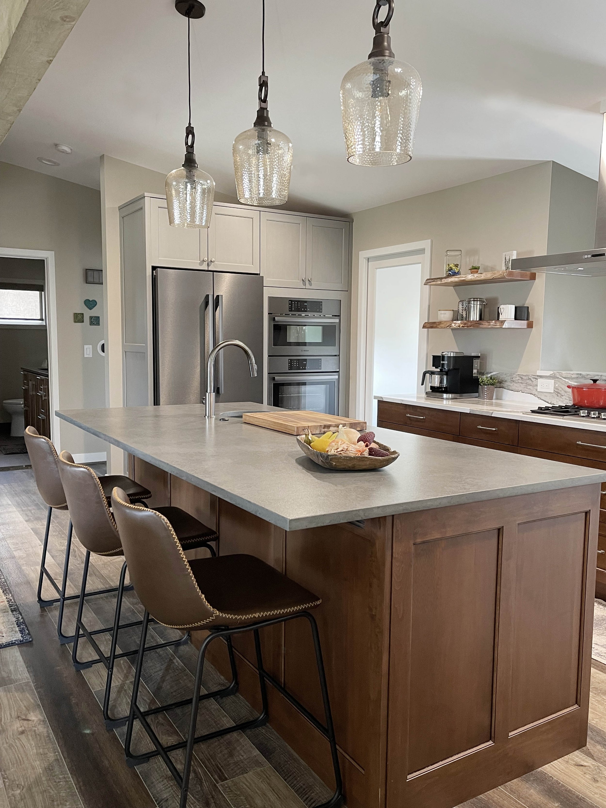 Modern kitchen with a large island island, three brown chairs with black legs, pendant lights, stainless steel appliances, and wooden shelves.