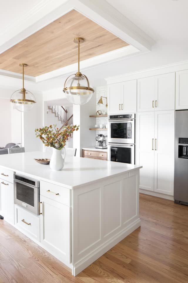 Modern white kitchen with an island, gold hardware, pendant lighting, wooden ceiling feature, stainless steel appliances, and wooden flooring.