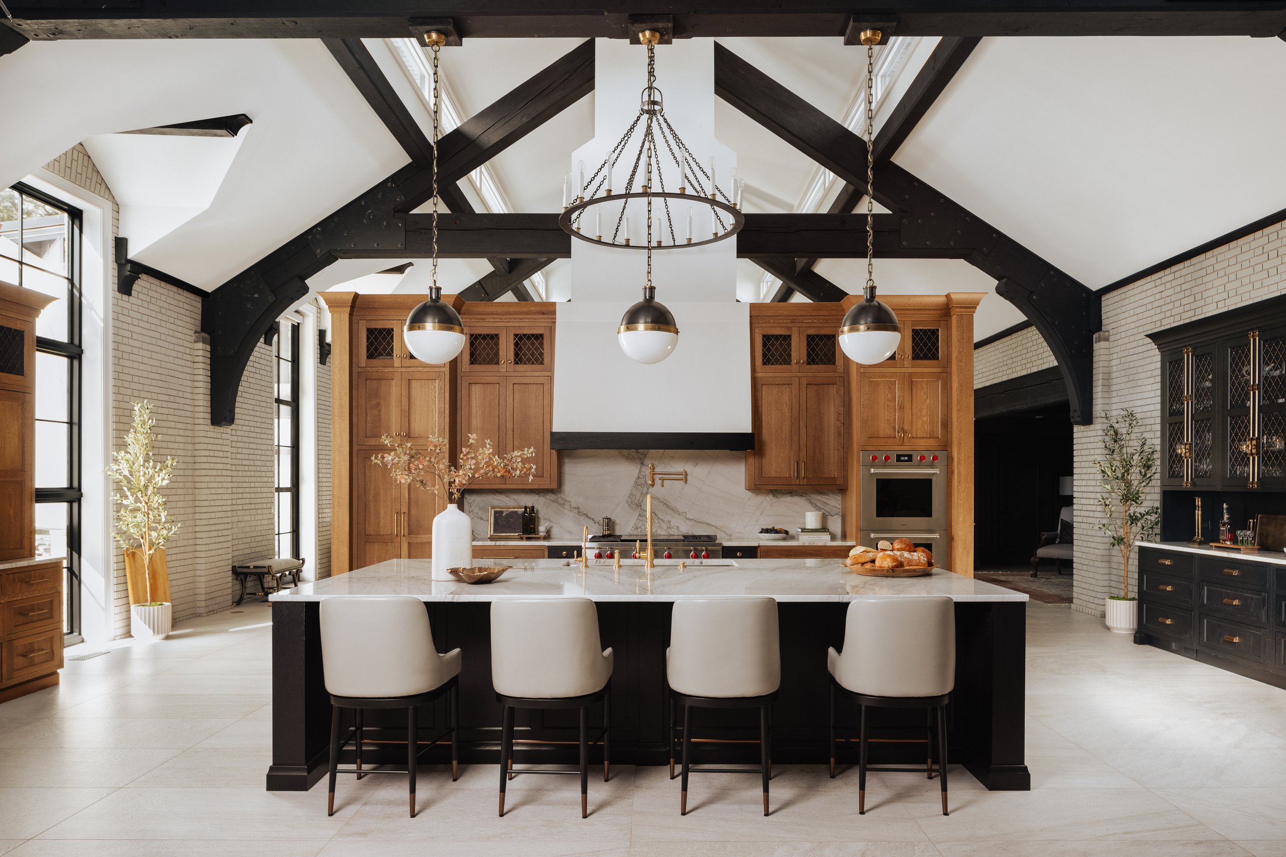 Modern kitchen with a large central island, beige chairs, wooden cabinets, black accents, high vaulted ceiling with black beams, pendant and chandelier lighting, large windows, and minimal decorative elements