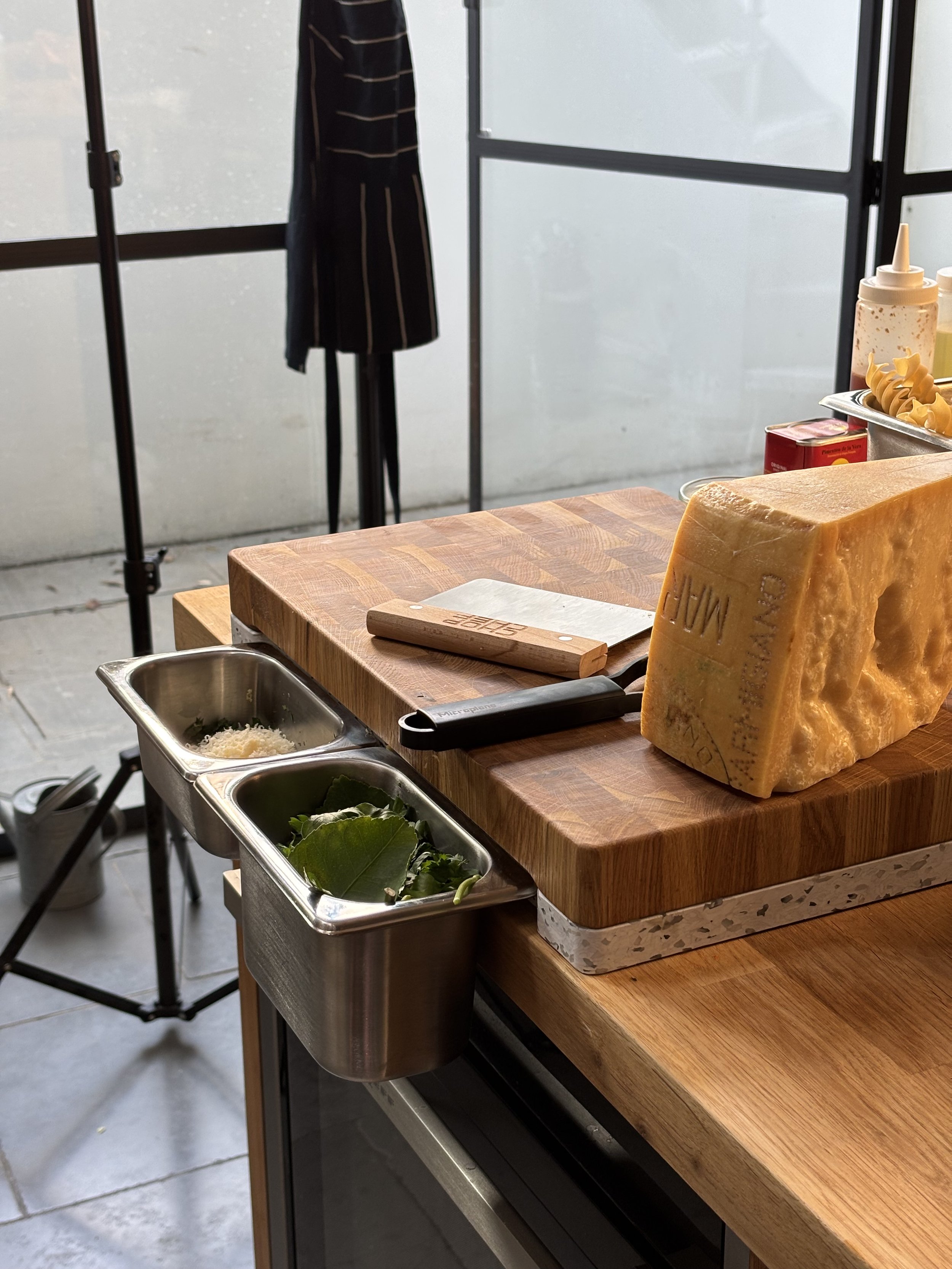 A cheese block on a wooden cutting board with cheese knives and fresh greens in a metal container. Behind is a counter with condiments and utensils, and a window with a mannequin dressed in a striped outfit in the background.