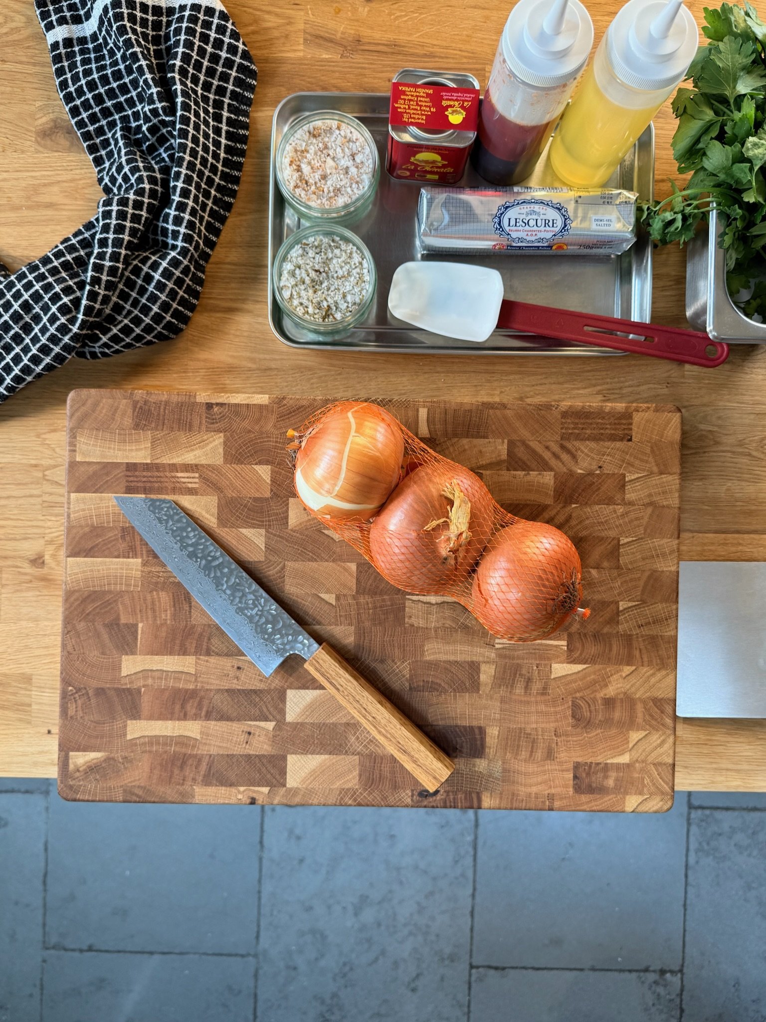 Three onions in a mesh bag on a wooden cutting board with a Japanese knife, set on a kitchen counter.