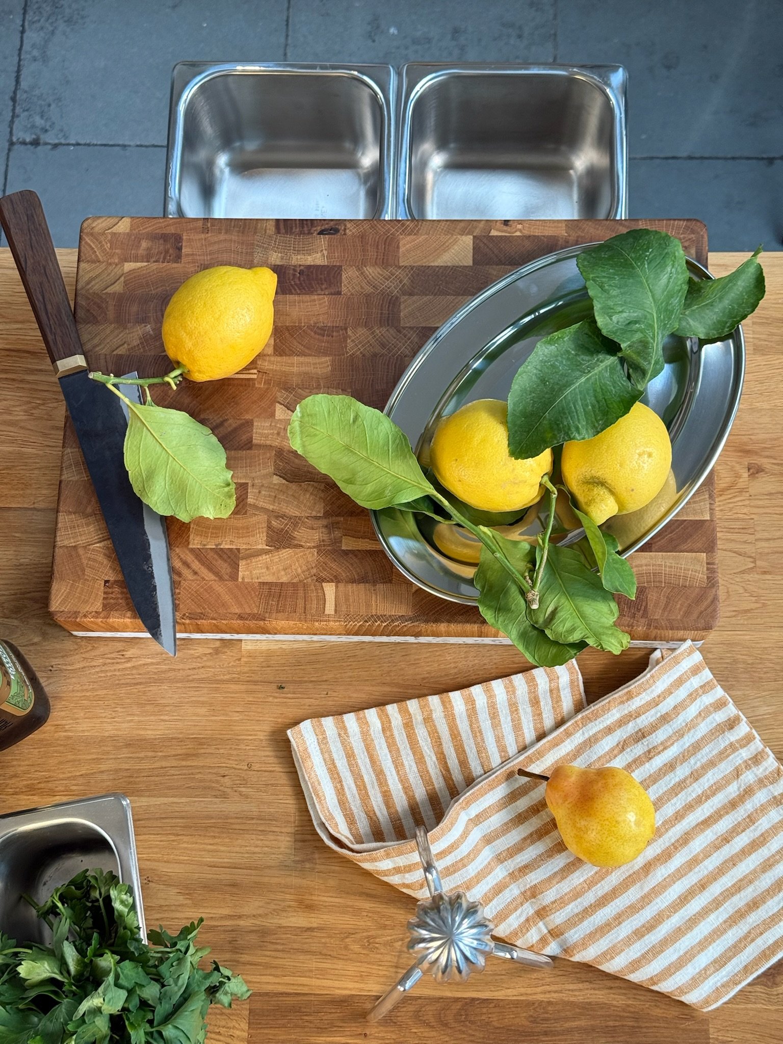 A wooden kitchen countertop with a cutting board, fresh lemons with leaves, a lemon squeezer, a striped cloth towel with a pear, and some metal containers in the background.