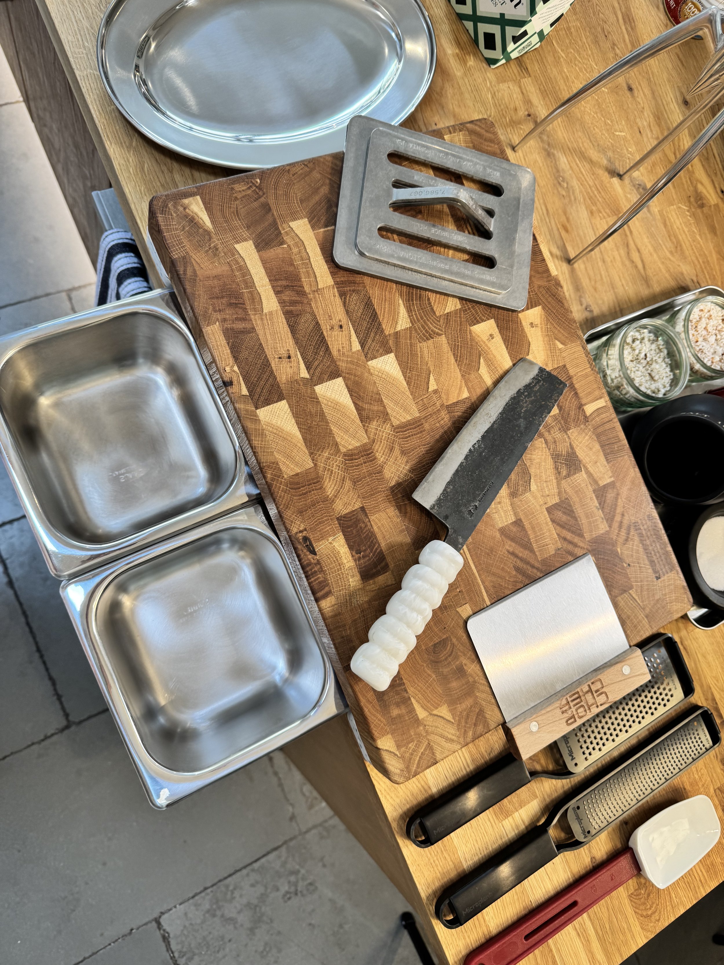 A wooden countertop with a cutting board, a cleaver, two metal molds, a metal scraper, a white plastic scraper, a cheese grater, and a white chopping tool. There are also metal containers, a metal tray, and jars in the background.