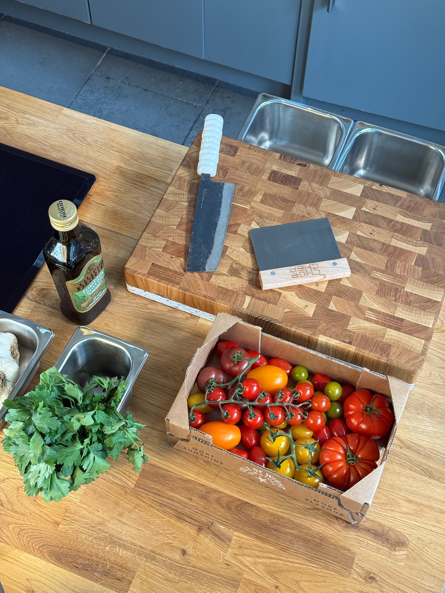 A wooden kitchen table holding a box of assorted heirloom tomatoes, a bunch of fresh parsley, and a bottle of balsamic vinegar. There is a bread knife with a white handle, a small black cutting board, and some kitchen knives on a wooden cutting board