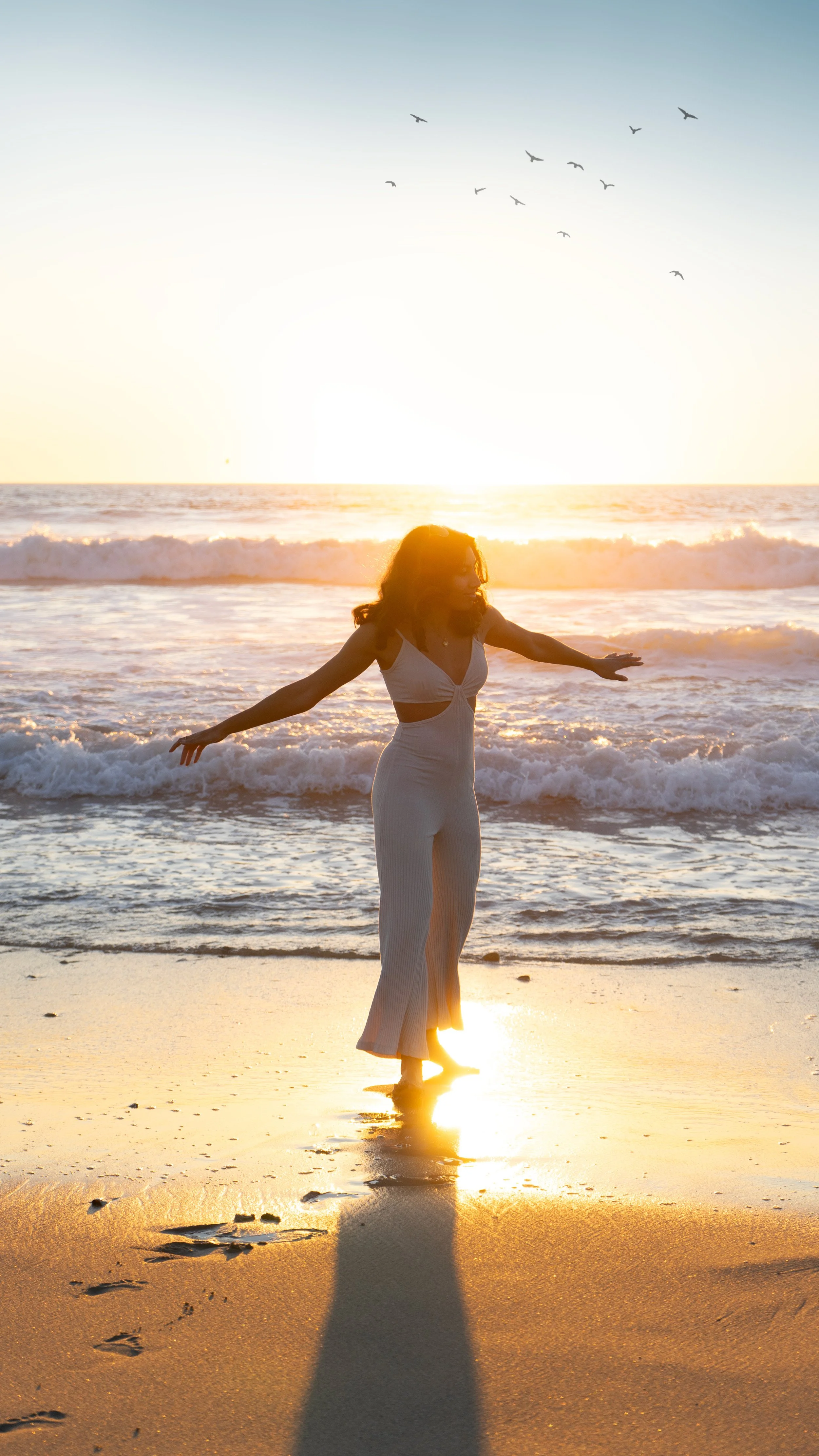 A woman in a white dress standing on the beach with arms outstretched at sunset, with the sun reflecting on the wet sand and waves, and birds flying in the sky.