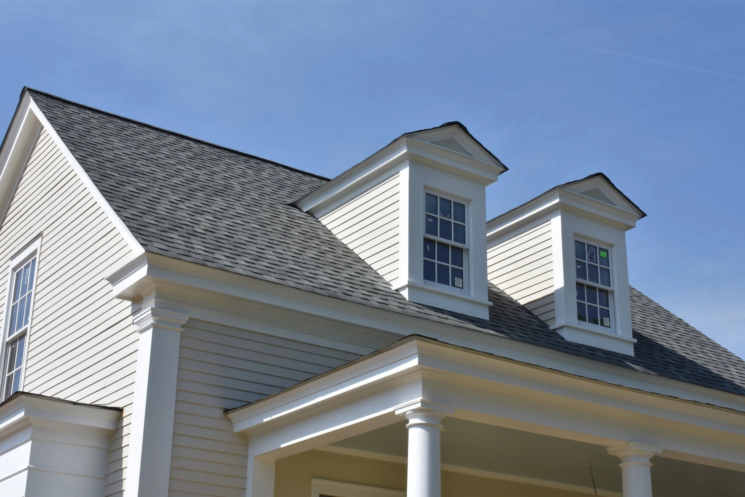 Close-up of a white house with traditional architecture, featuring gabled dormer windows, white siding, and a gray shingle roof against a blue sky.