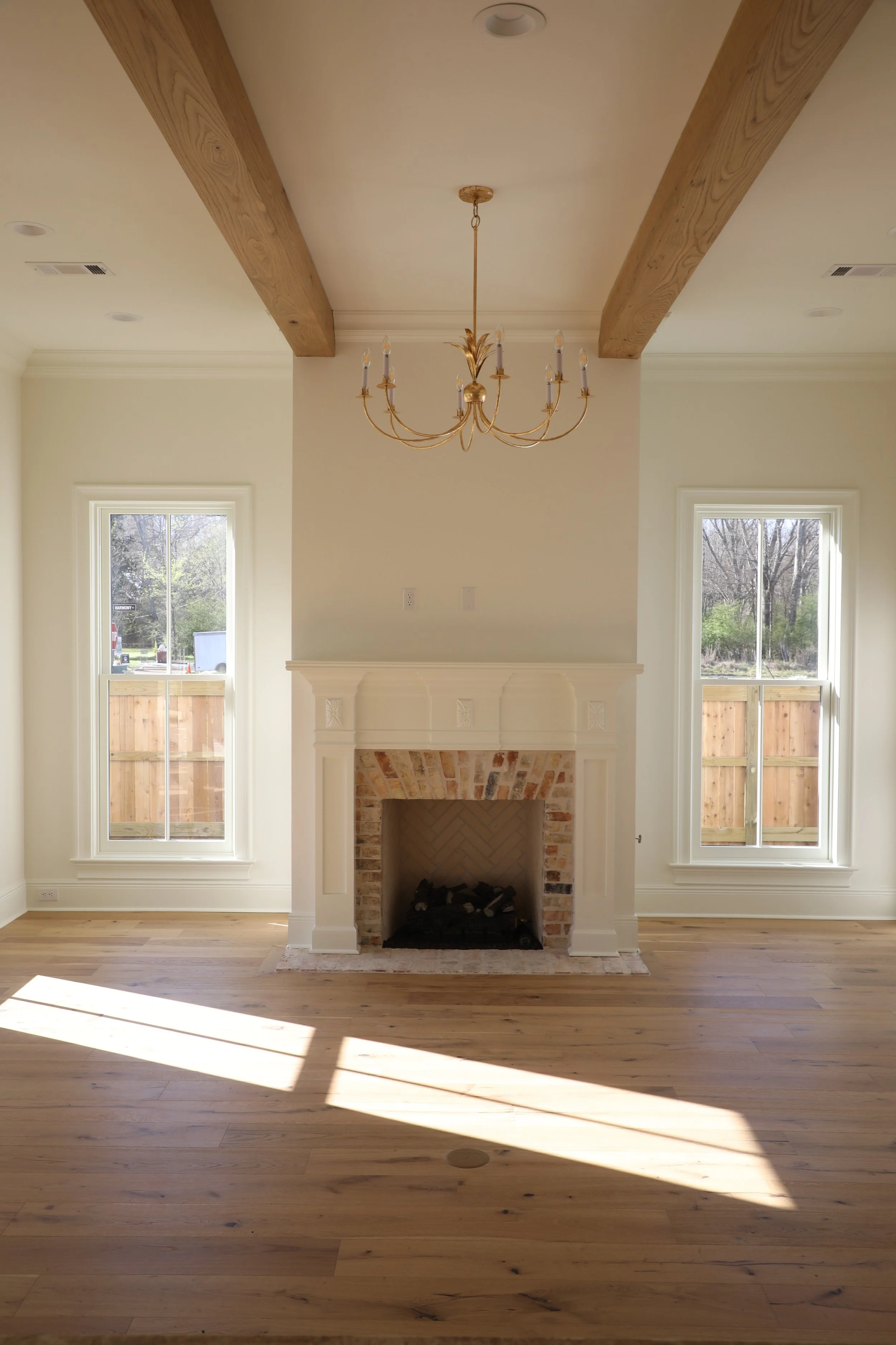 Living room with a white brick fireplace, two large windows with wooden fences outside, a gold chandelier, and wooden beams on the ceiling, with hardwood floors.