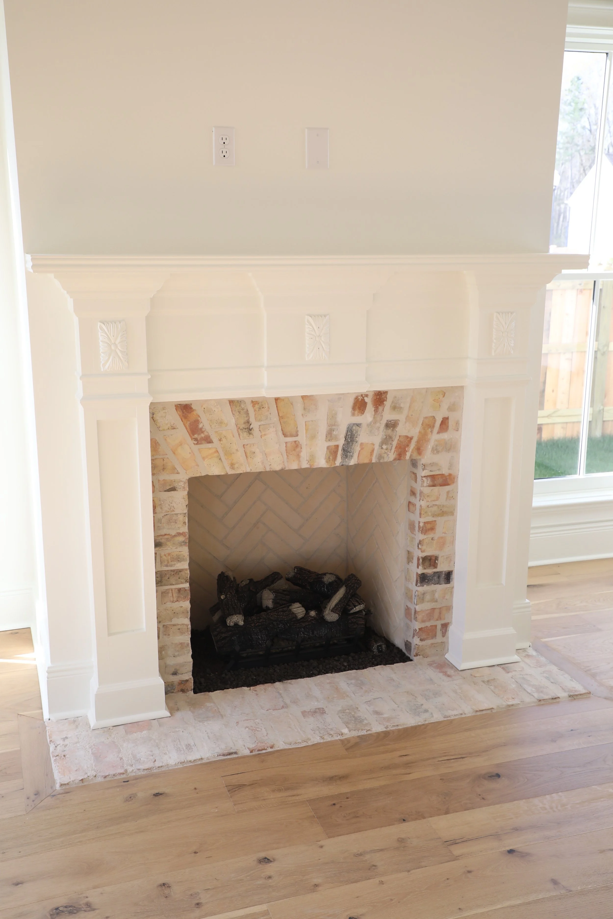 A white fireplace with decorative woodwork and brick interior, with logs inside, situated on a wooden floor near a glass door.
