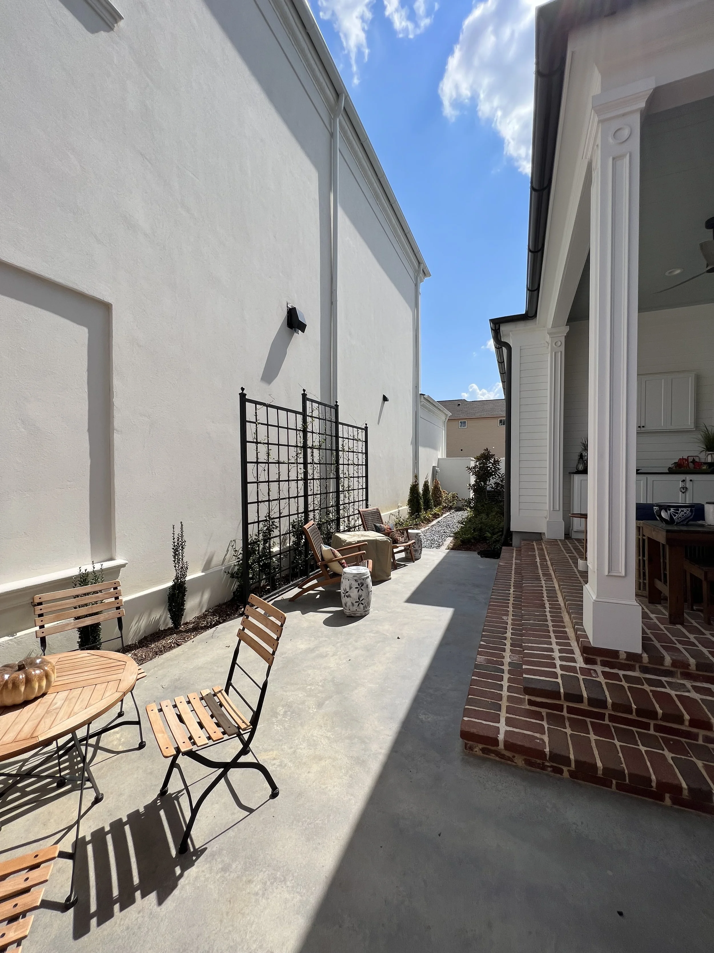 A backyard patio with a small round table and chairs, a sitting area with a bench and a small table, and a garden bed with plants, under a bright blue sky with clouds.