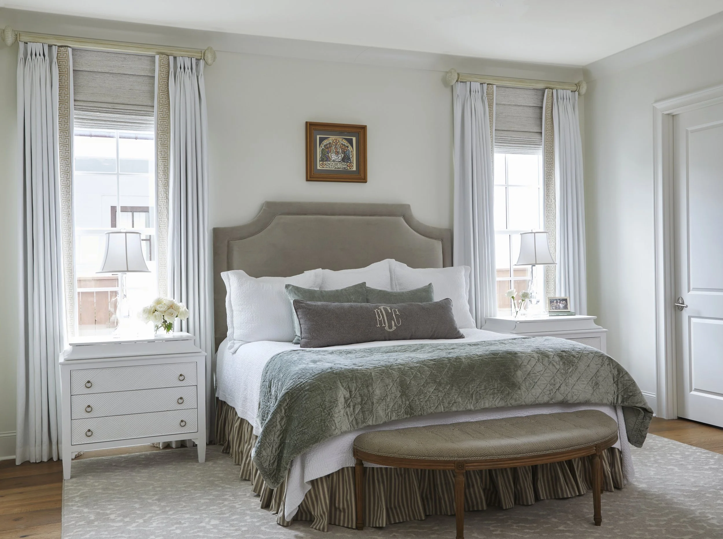 A cozy bedroom with a large bed featuring a beige headboard, white and gray bedding, and decorative pillows. Flanked by two white nightstands with lamps, flowers, and photo frames. Two windows with white curtains and beige blinds, and a framed pictur