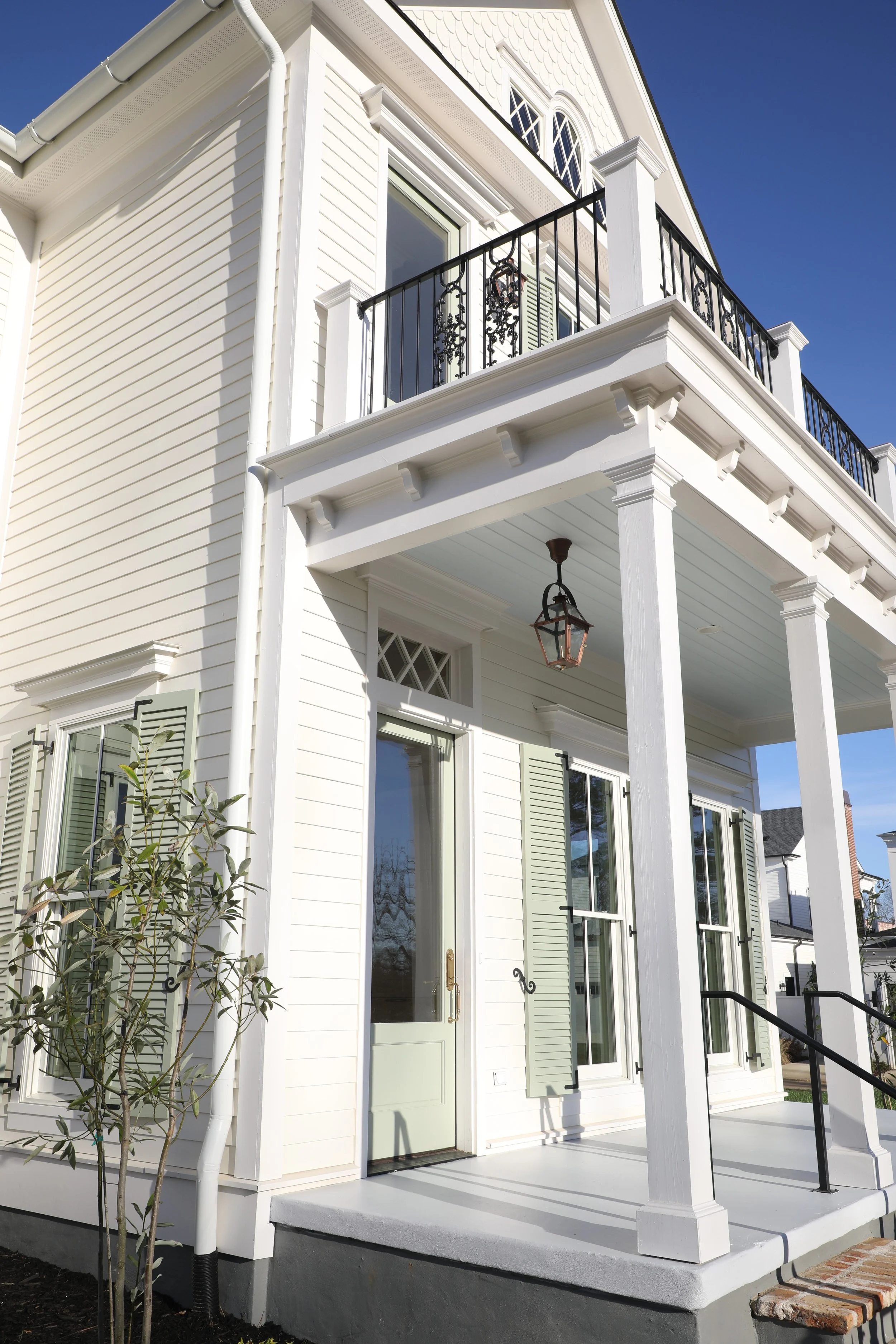 Side view of a modern white house with green shutters, a small front porch with columns, and a balcony with a decorative black railing and a hanging lantern.