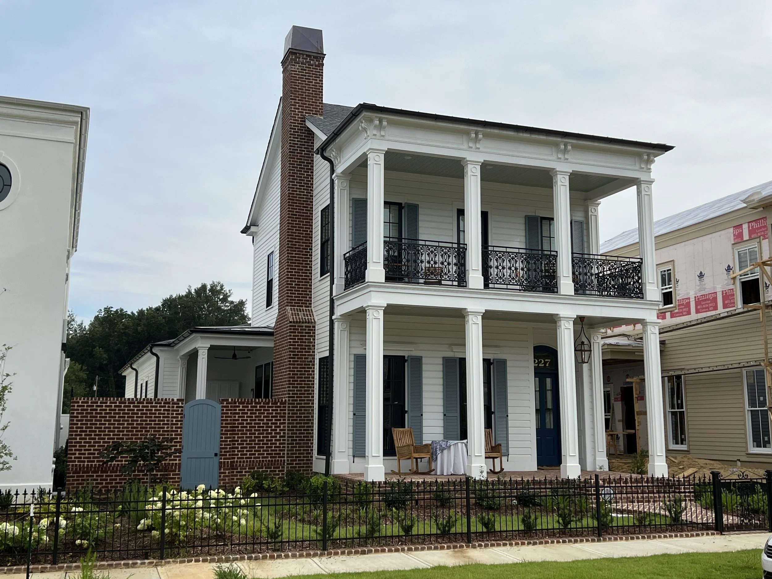 A two-story white house with blue shutters, a porch with balcony, black wrought iron railings, and a brick chimney. There is a small garden with a black fence in front.