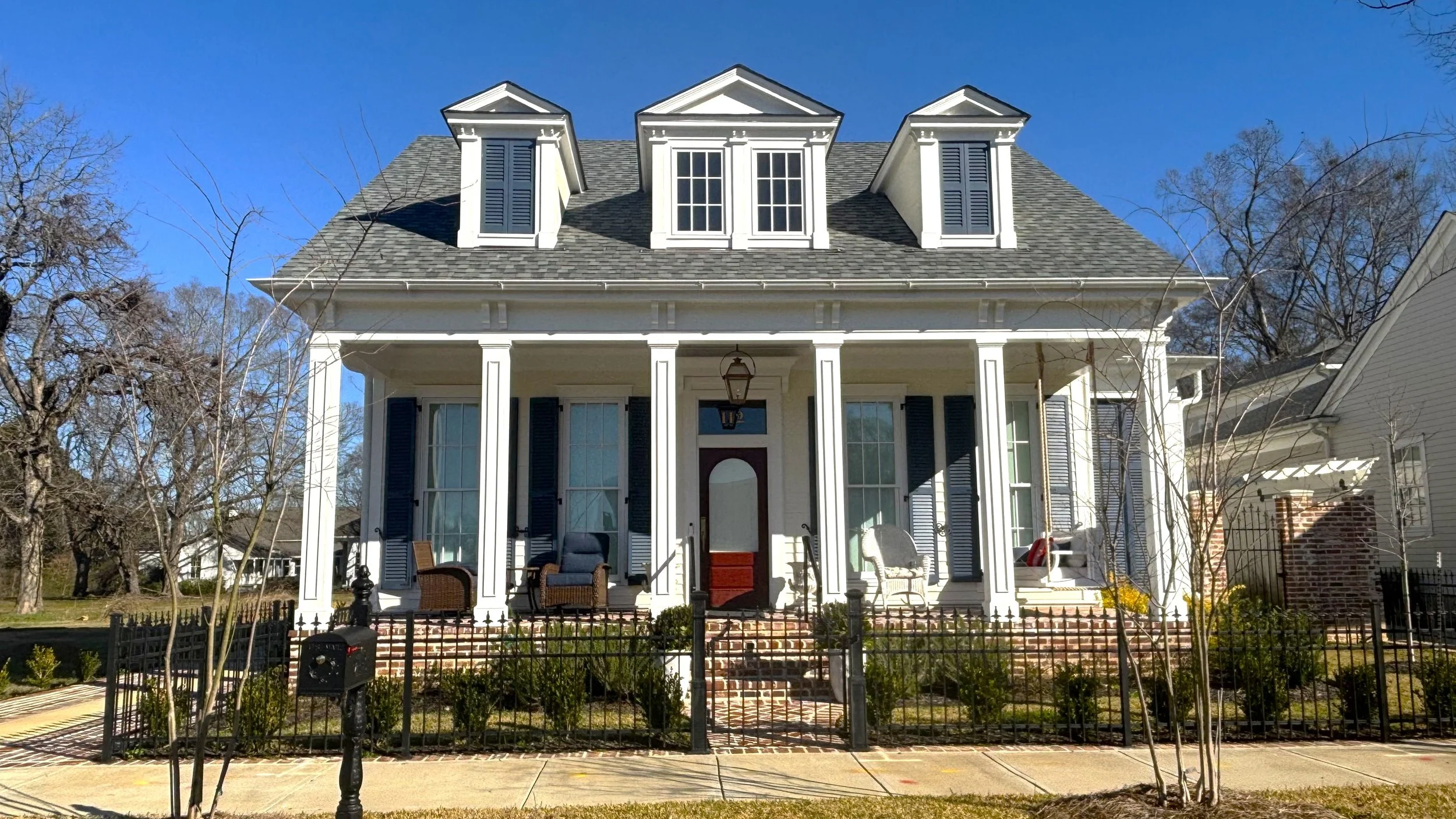 A large, white, two-story house with a front porch, blue shutters, and dormer windows on a sunny day.