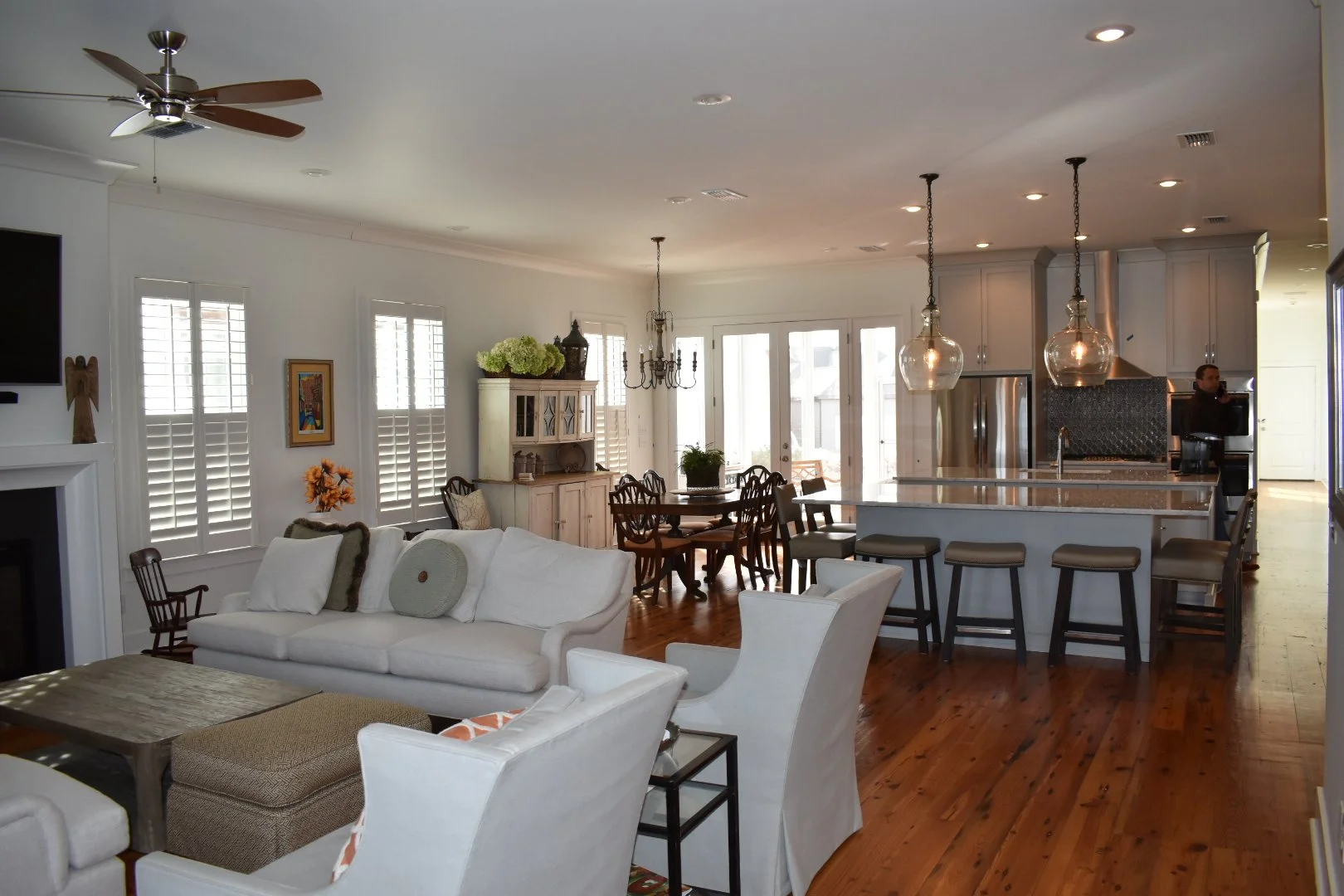Open-concept living room and kitchen with white walls, hardwood floors, white and beige furniture, bar stools, kitchen island, chandelier, ceiling fan, and large windows with shutters.