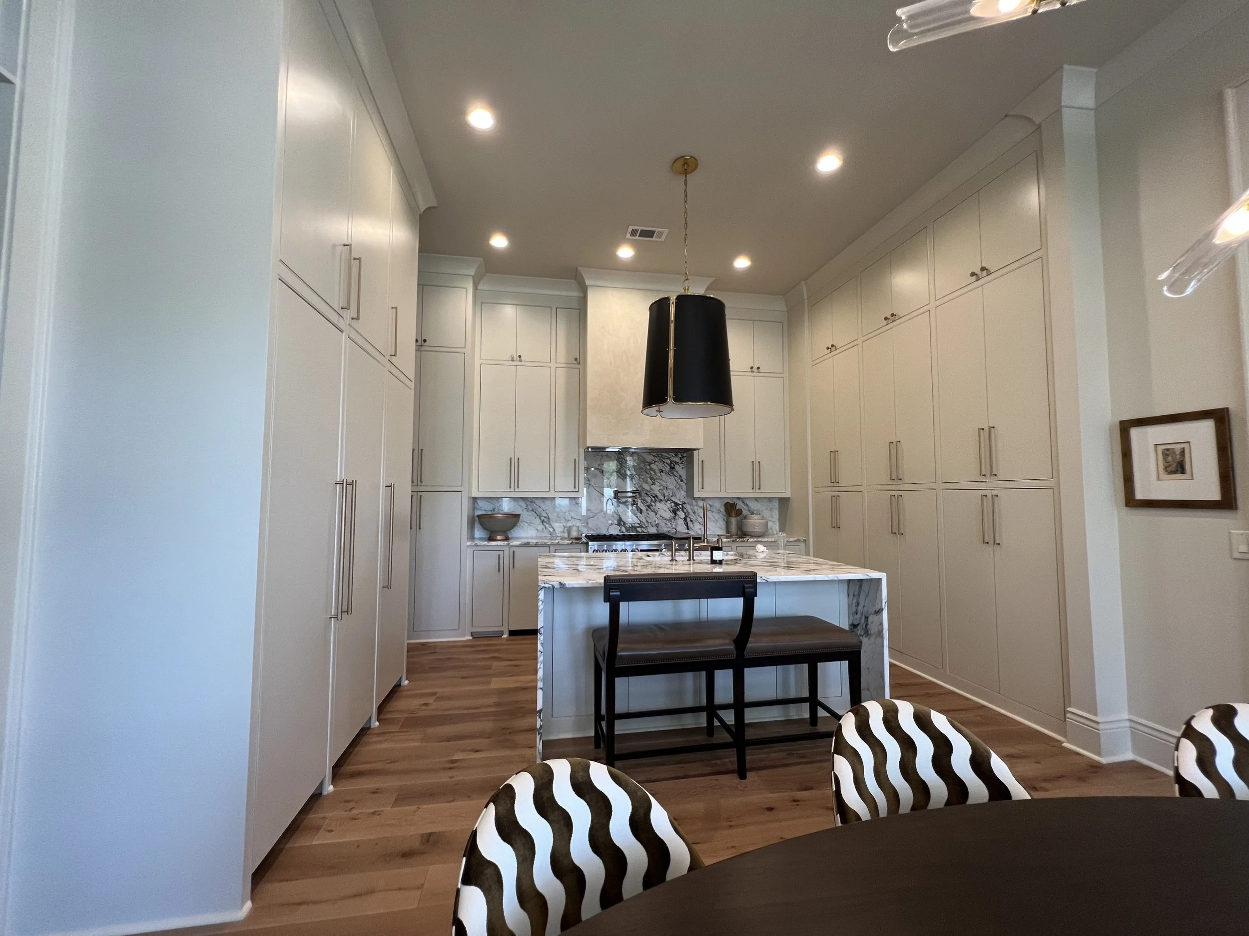 Modern kitchen with white cabinetry, marble countertops, and a black pendant light over a center island. Wooden flooring and a round dining table with patterned chairs are visible.