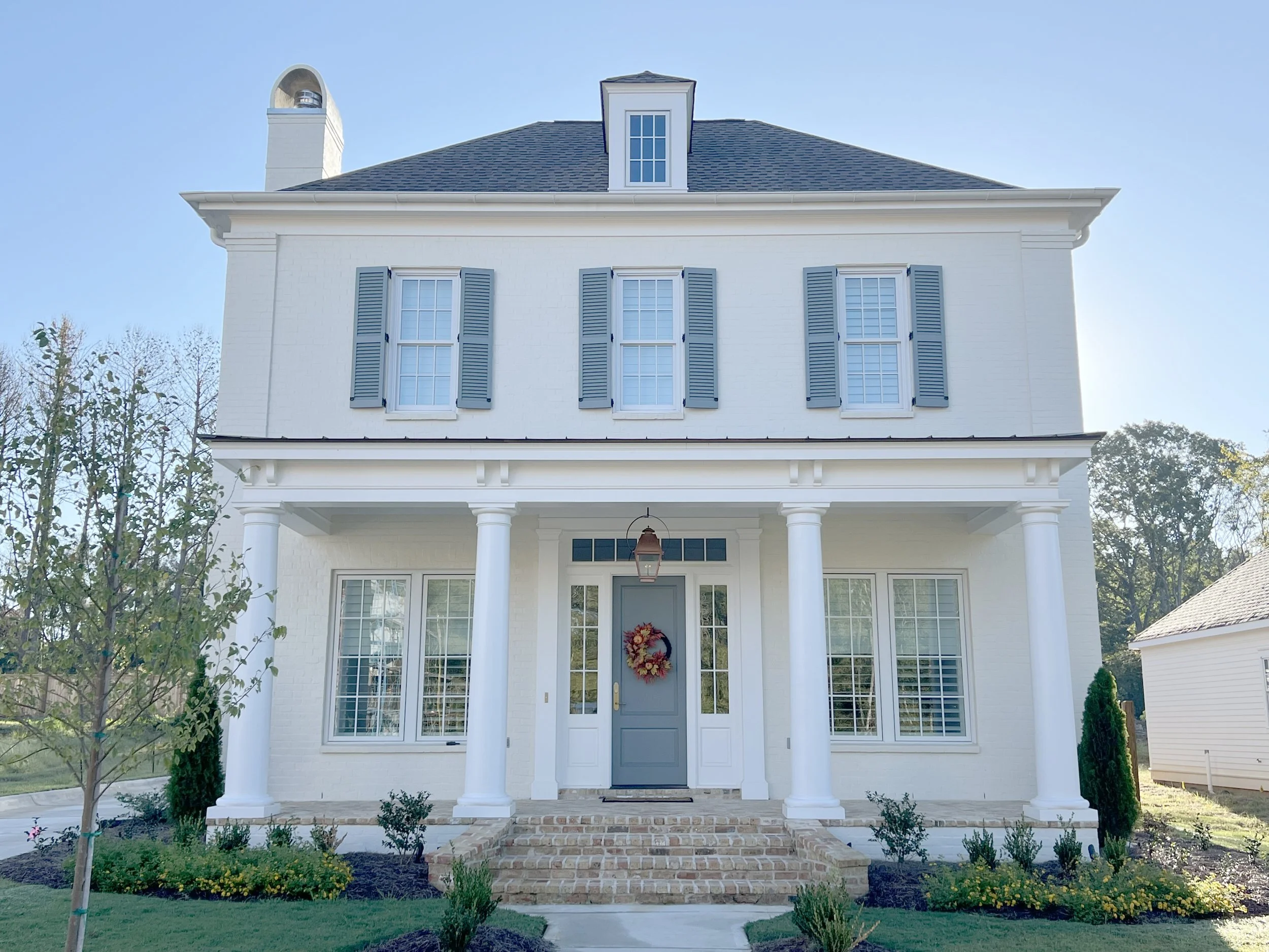 White two-story house with blue shutters, a porch with white columns, and a front door decorated with a floral wreath, set against a clear blue sky.