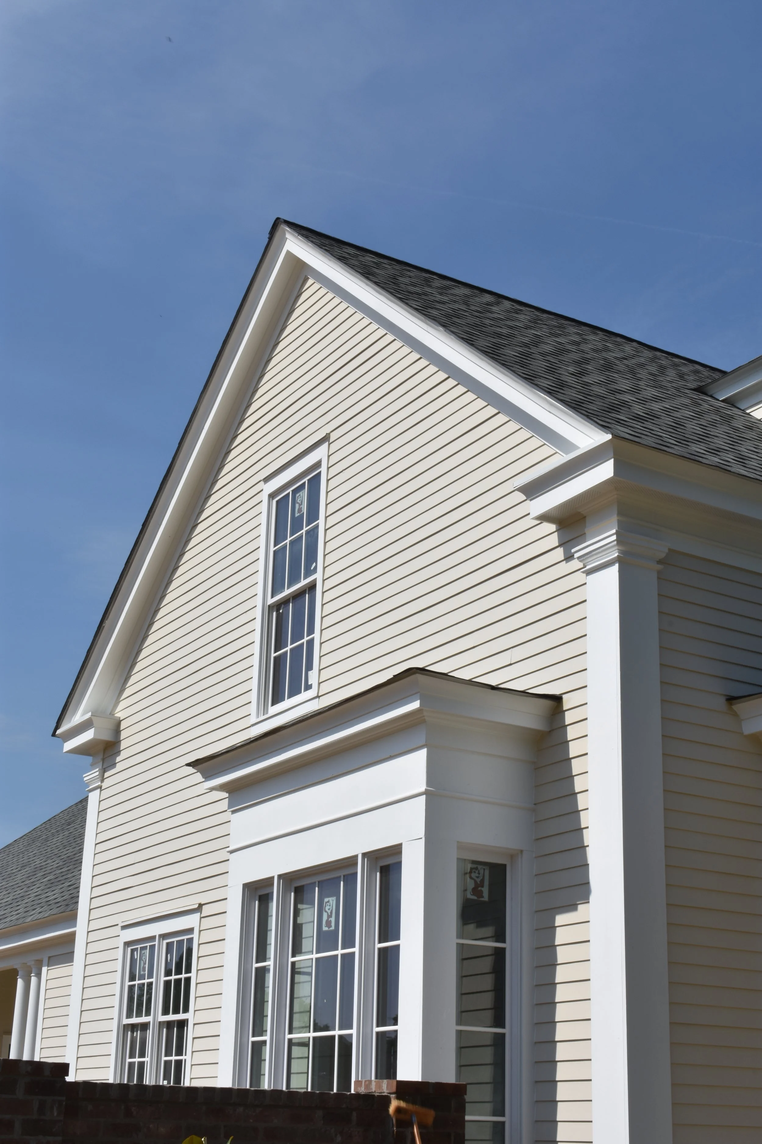 Close-up of a beige house exterior with white trim, double-hung windows, and a sloped roof under a clear blue sky.